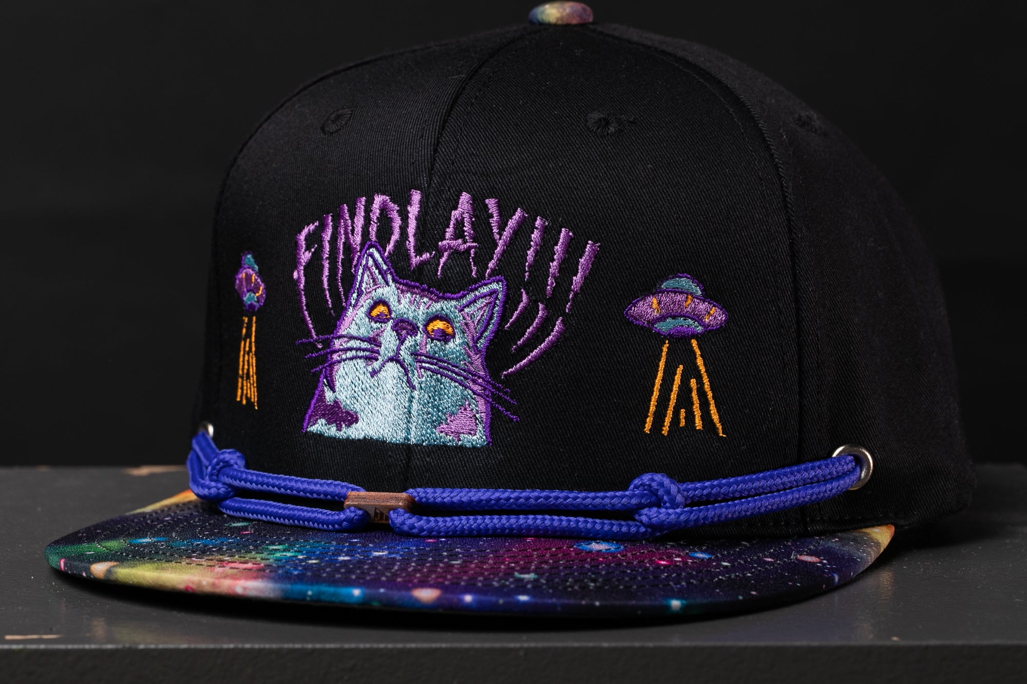 A black baseball cap with a colorful, space-themed design, including a cat and two alien-like figures.