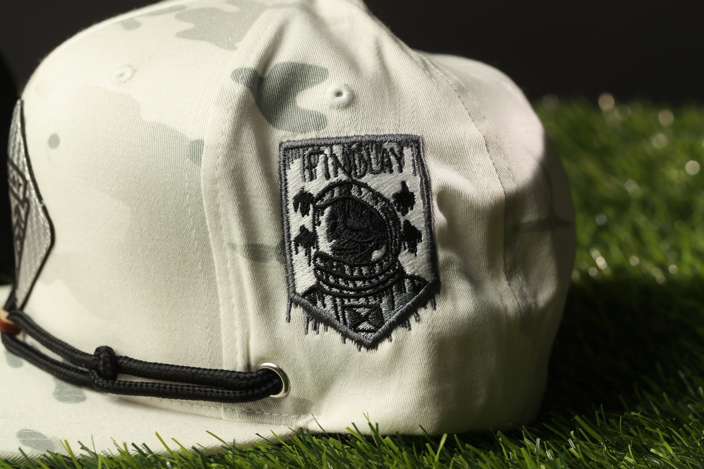 A white baseball cap with a black embroidered patch on the front, sitting on a grassy surface.