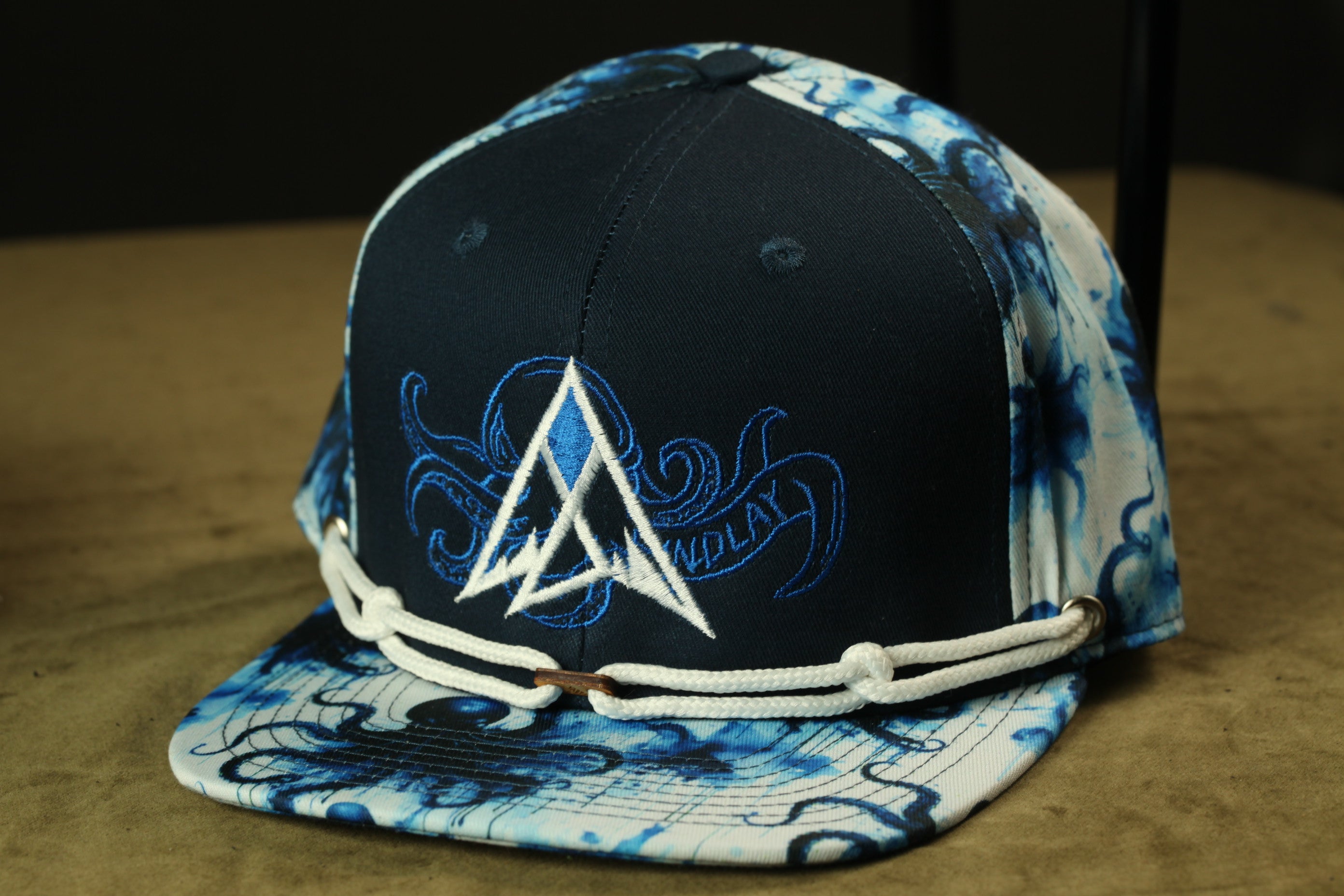 A black baseball cap with a blue and white patterned design, featuring a white triangle logo and a wave-like pattern.