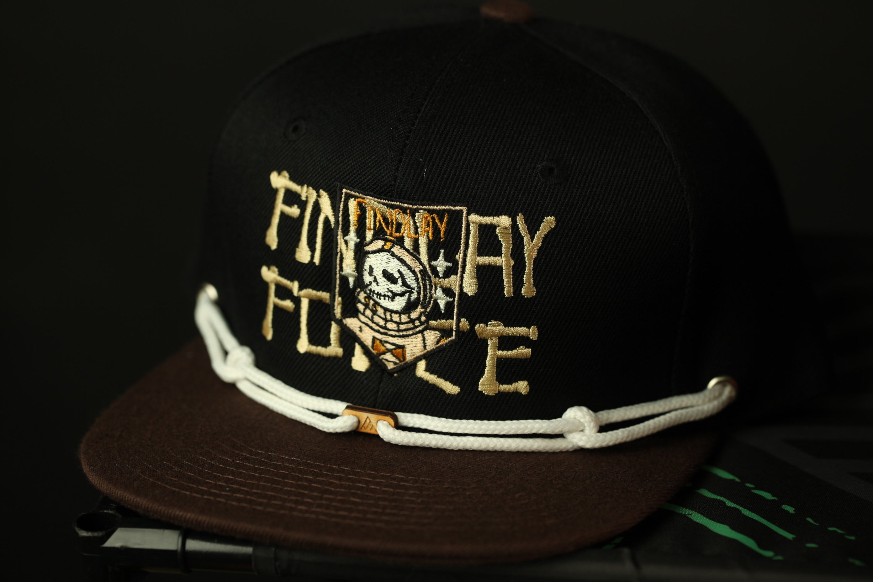 A black baseball cap with white and gold embroidered text and a skull design.
