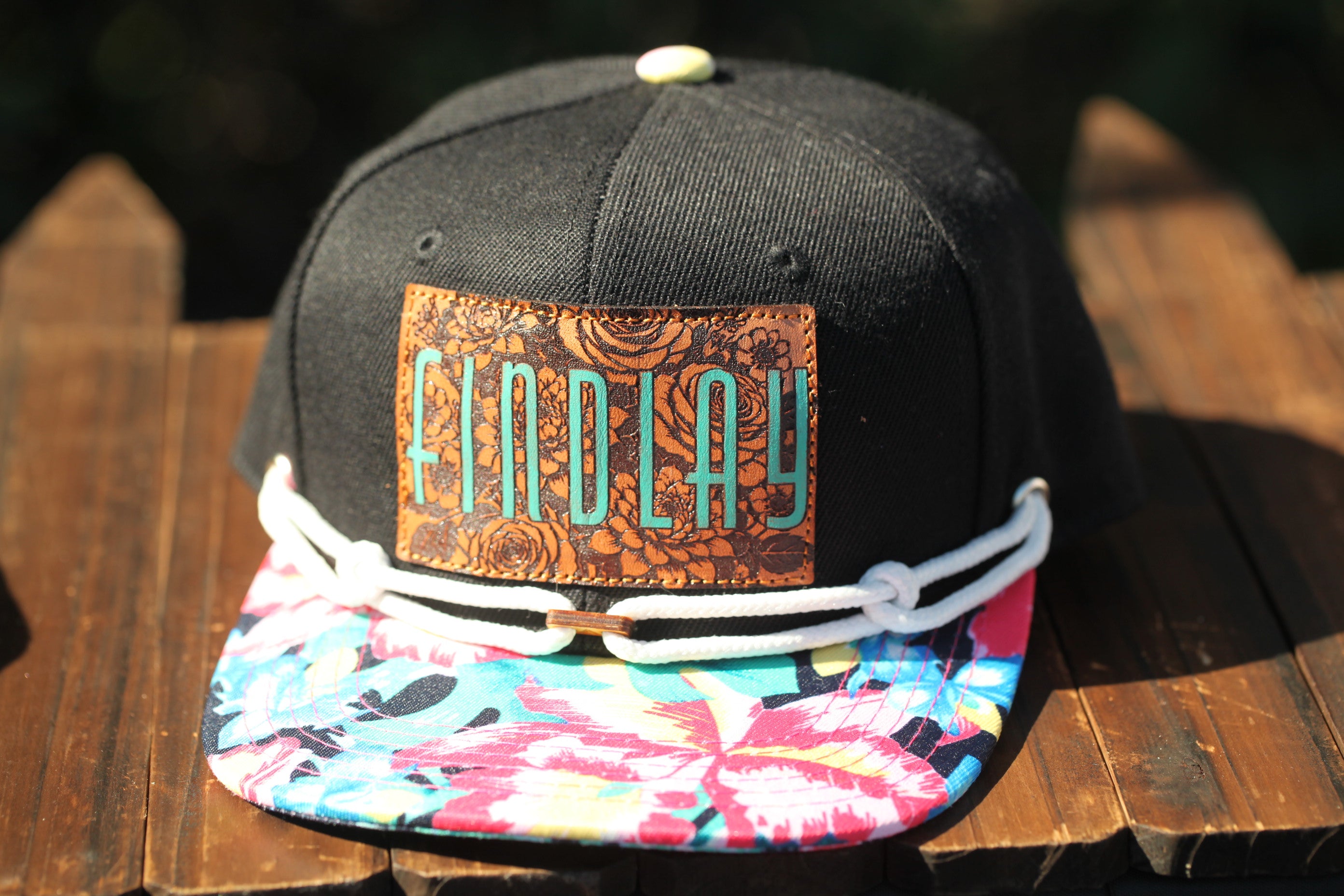 A black baseball cap with a floral pattern and the word "FINDlay" printed on it, sitting on a wooden surface.