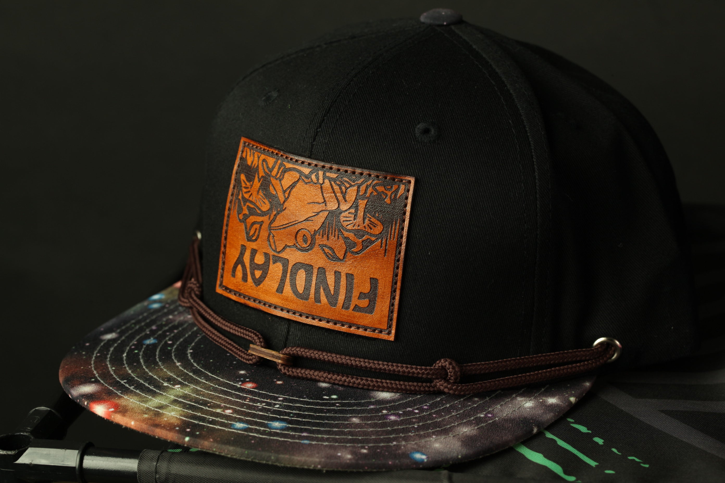 A black baseball cap with a colorful galaxy print and a leather patch on the front.