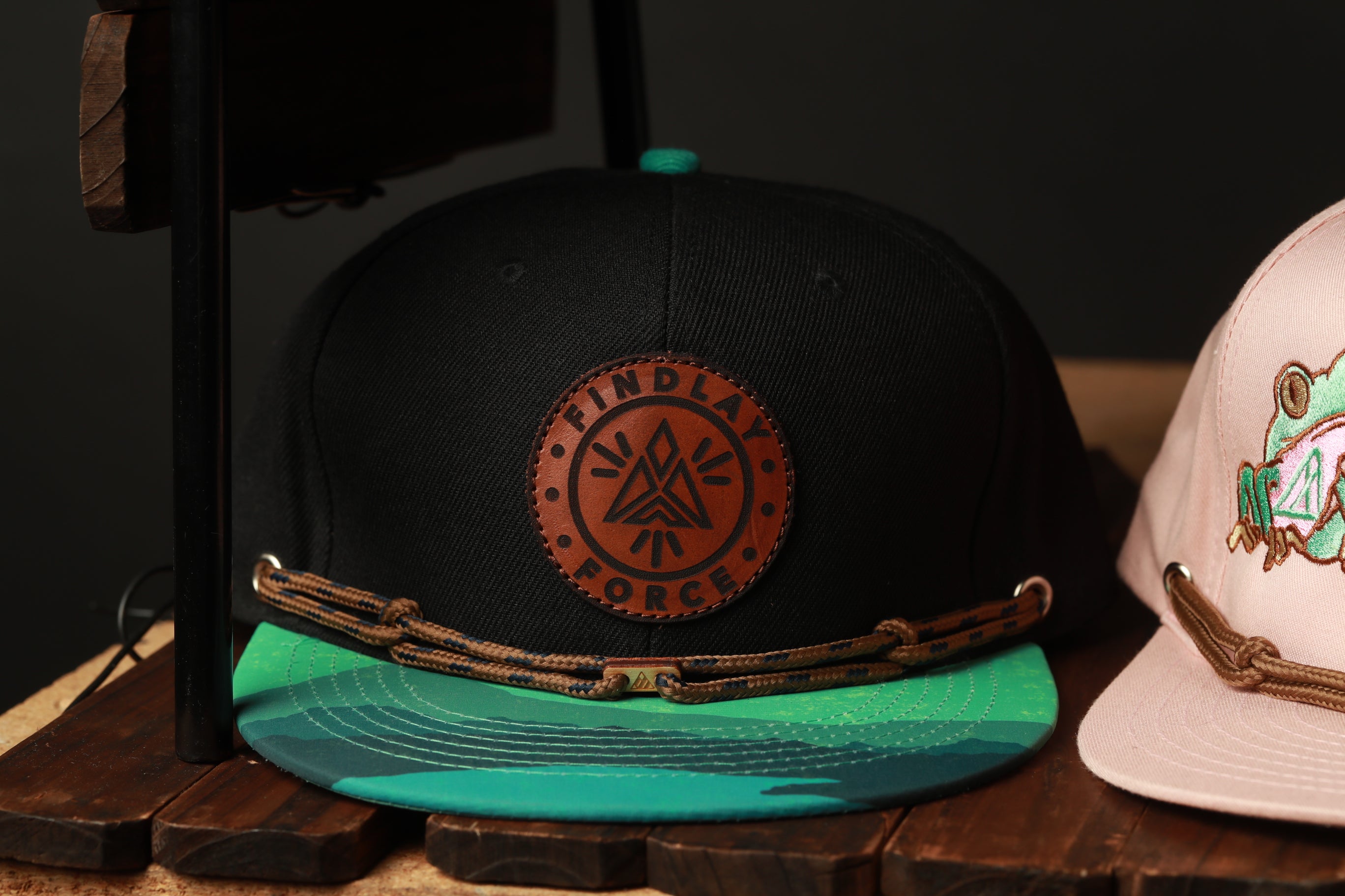 A black baseball cap with a circular logo and a green brim, placed on a wooden surface next to a pink baseball cap.