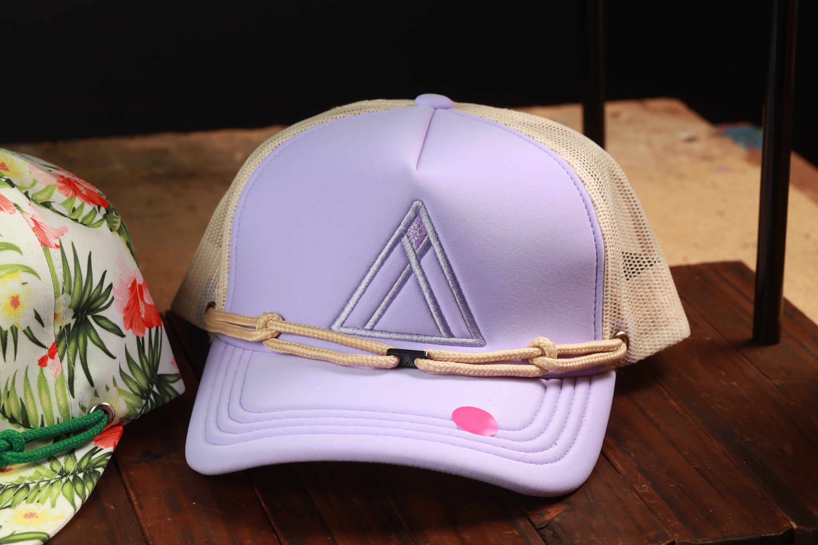 A purple baseball cap with a mesh back and a beige strap is displayed on a wooden surface, next to a floral print cap.