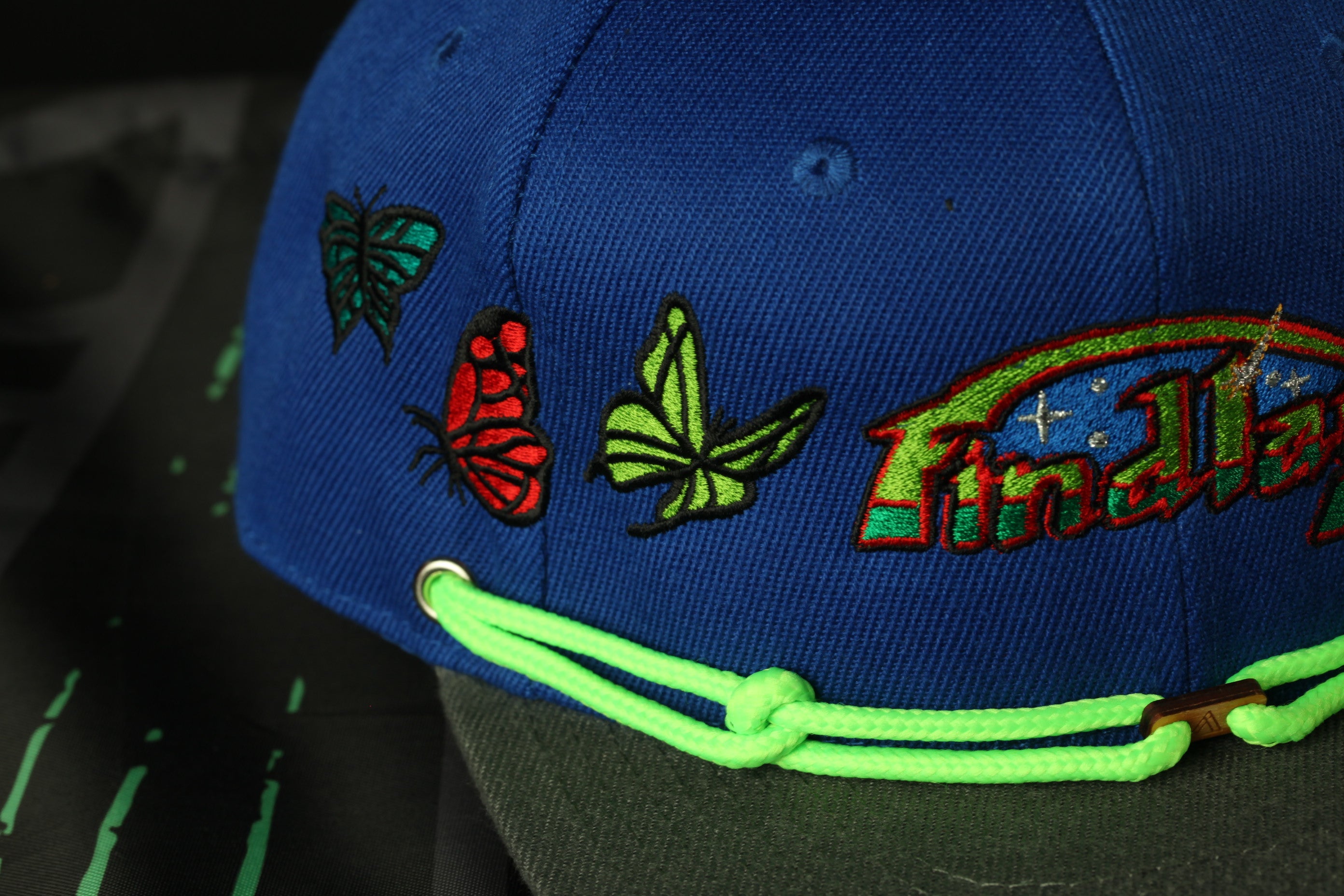 A close-up view of a blue baseball cap with colorful butterfly and flower embroidery, and a neon green string around the brim.