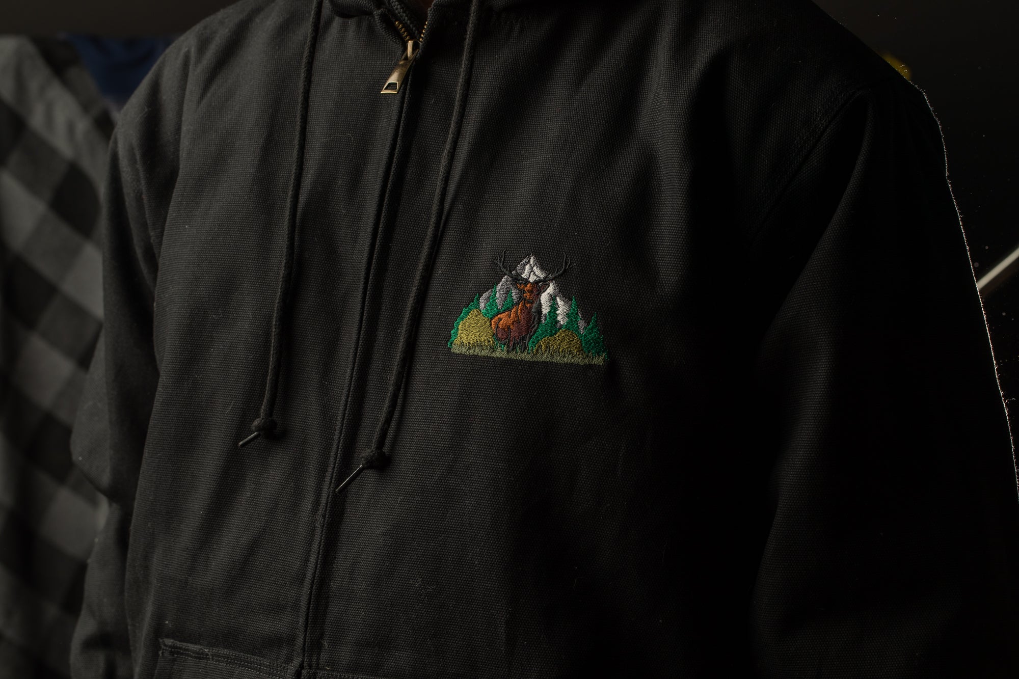 A close-up view of a person wearing a black hooded jacket with a small embroidered mountain scene on the front.