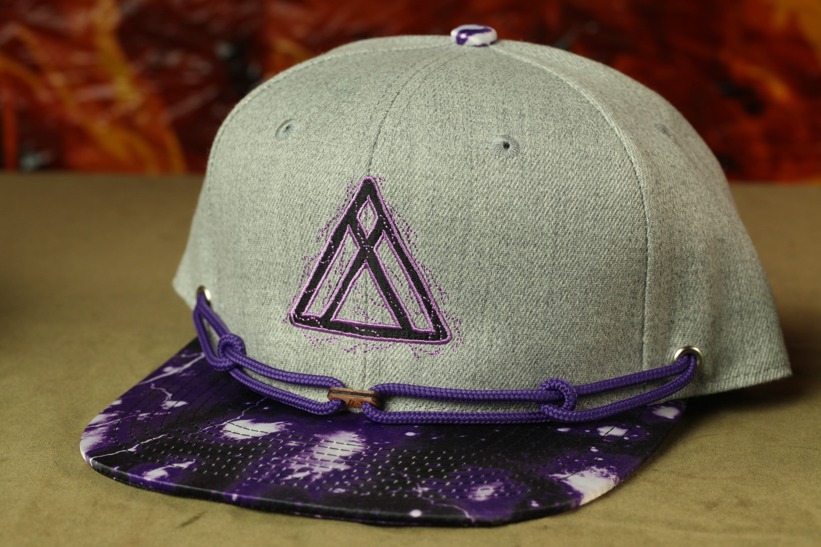 A gray baseball cap with a purple and white tie-dye pattern and a black triangle logo on the front.