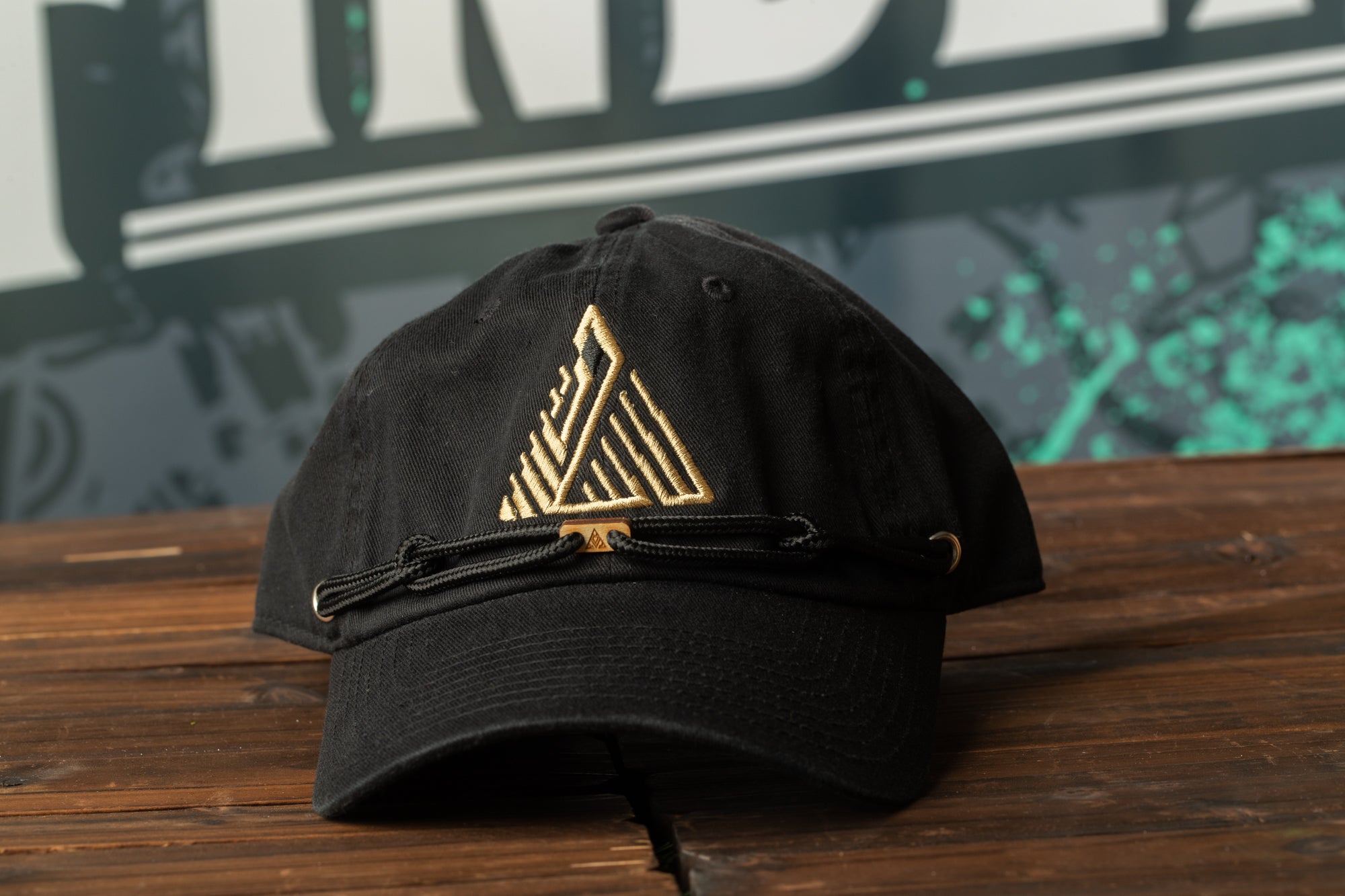 A black baseball cap with a gold geometric pattern on the front, placed on a wooden surface.