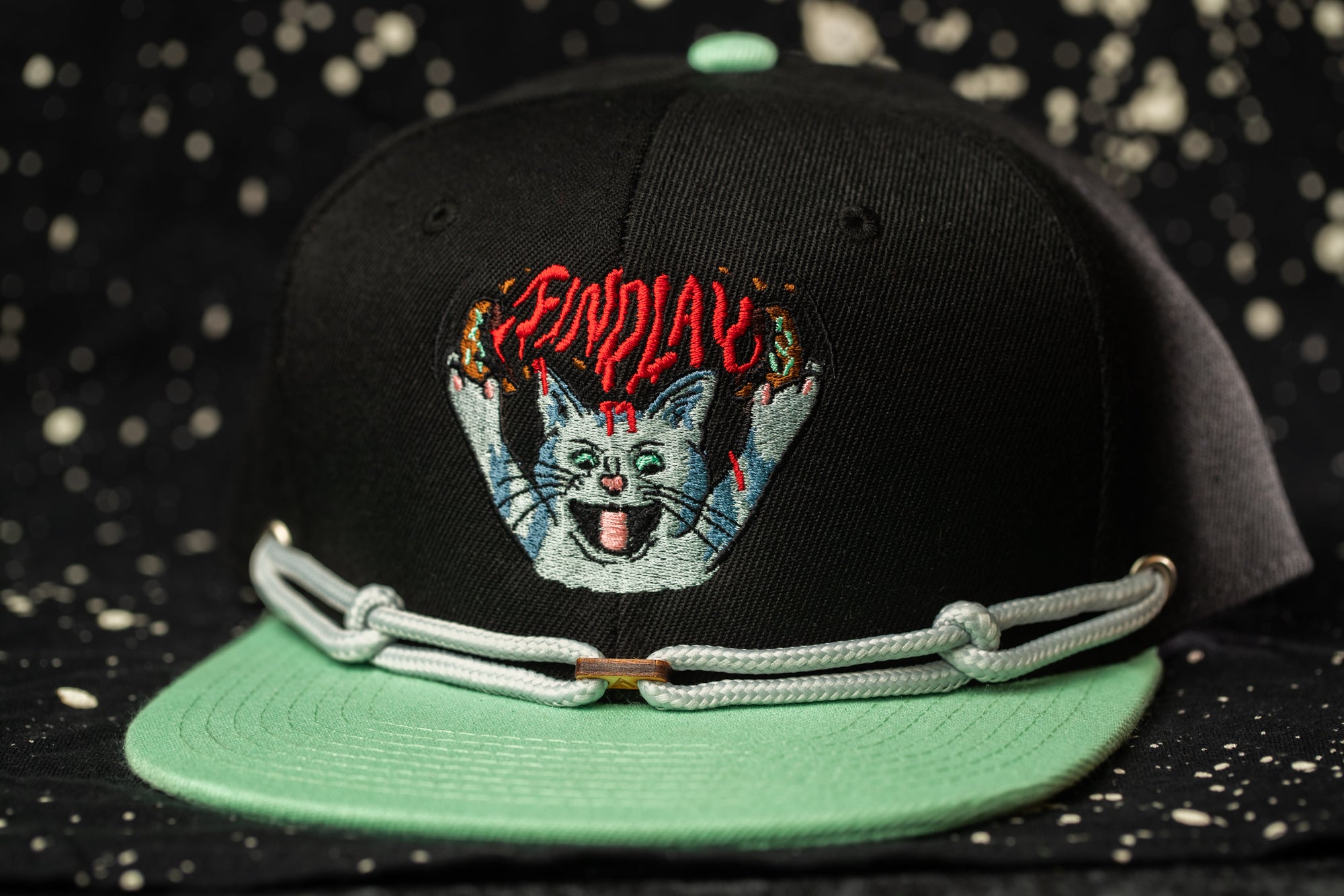 A black baseball cap with a green brim and a cat embroidered on the front, set against a black background with white speckles.