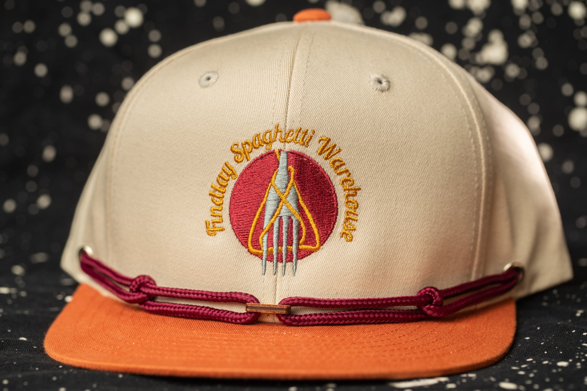 A beige baseball cap with a maroon band and a logo on the front, against a black background with white speckles.