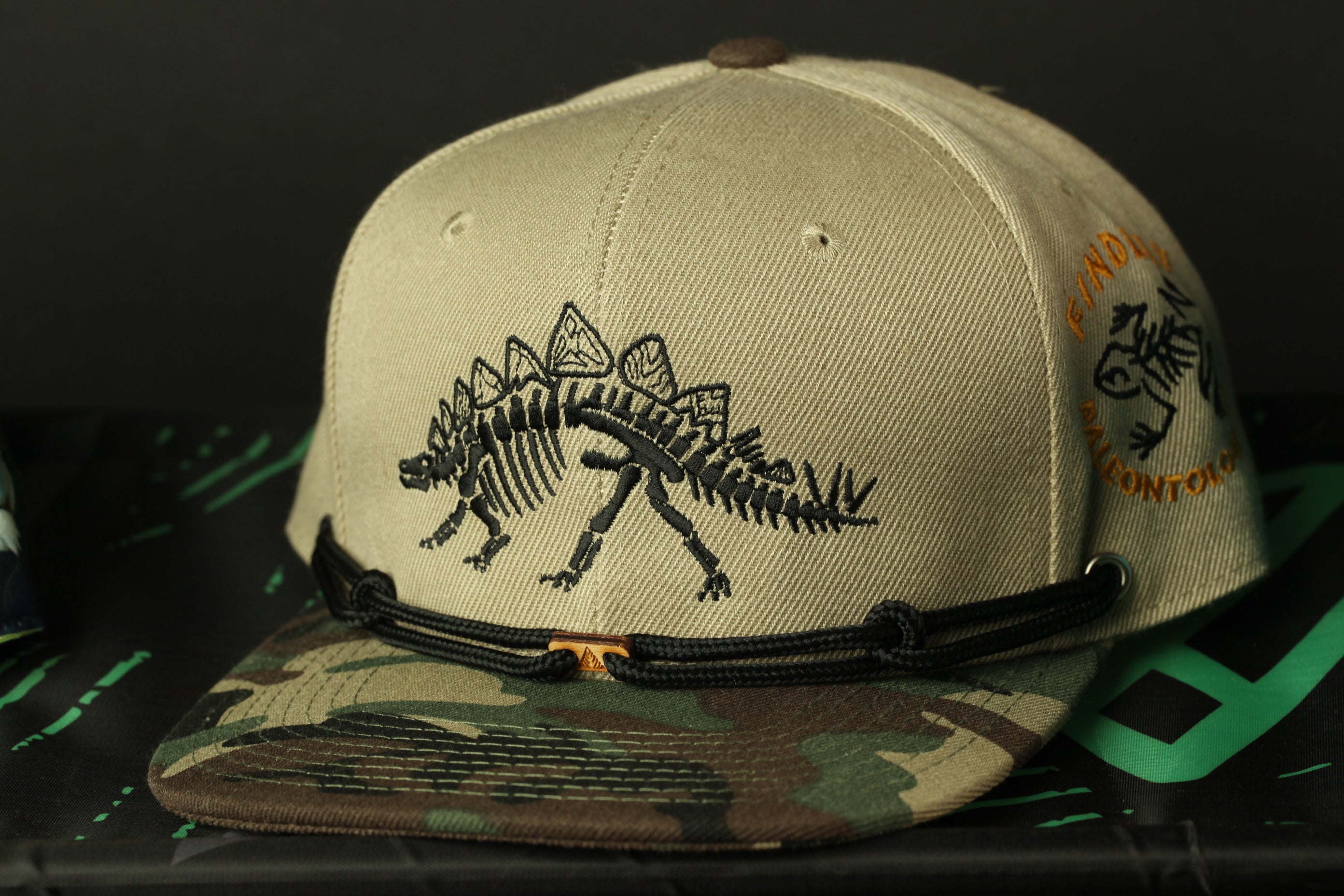A beige baseball cap with a dinosaur design on the front and a camouflage pattern on the brim, placed on a black surface.