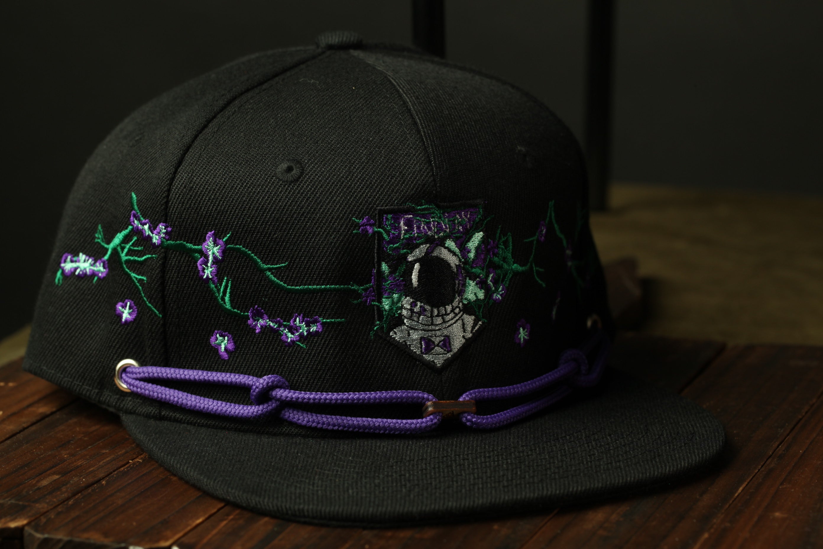 A black baseball cap with purple and green embroidered designs, including a small astronaut figure, is displayed on a wooden surface.