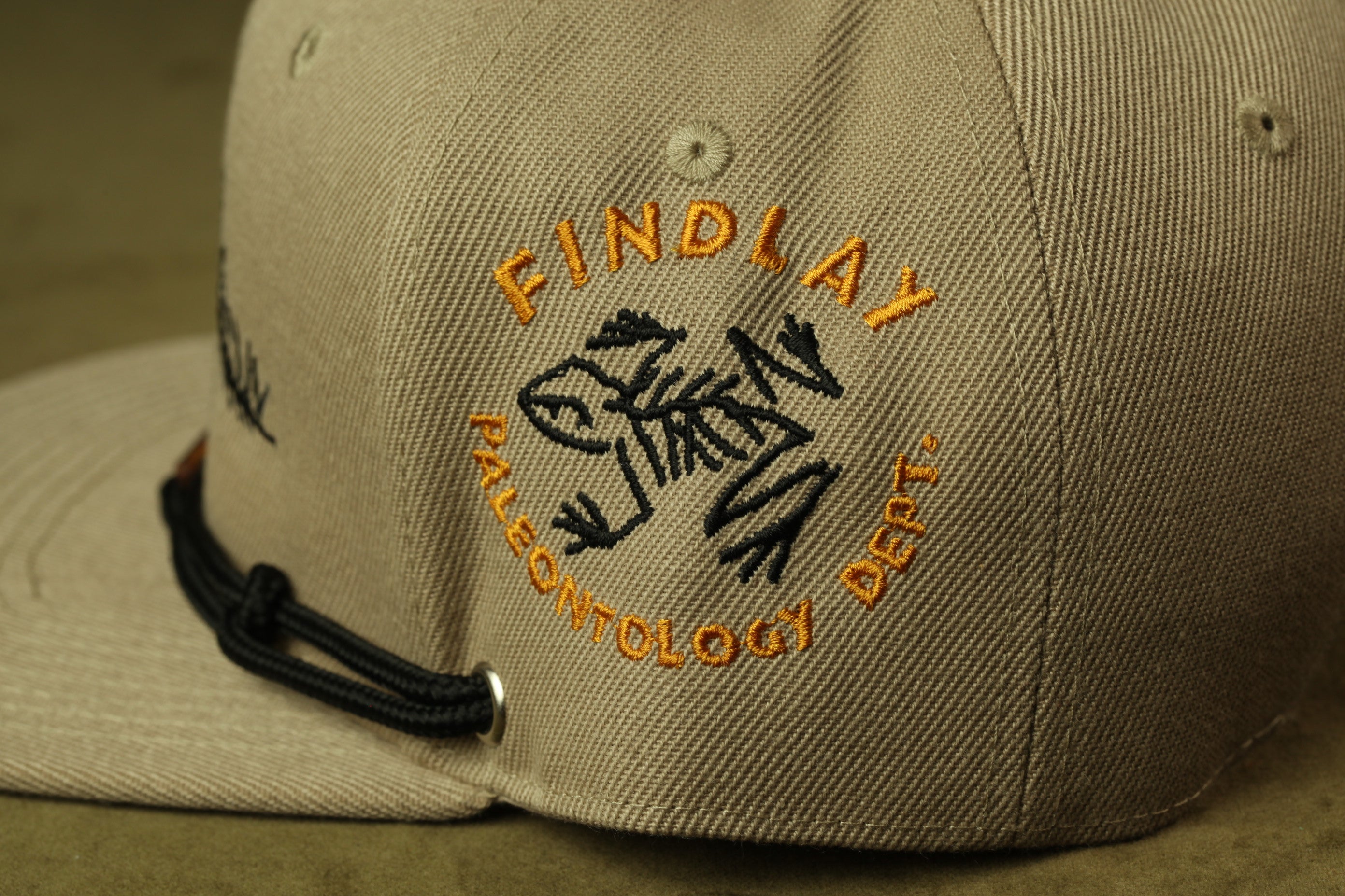 A close-up view of a beige baseball cap with a black and orange logo on the front.