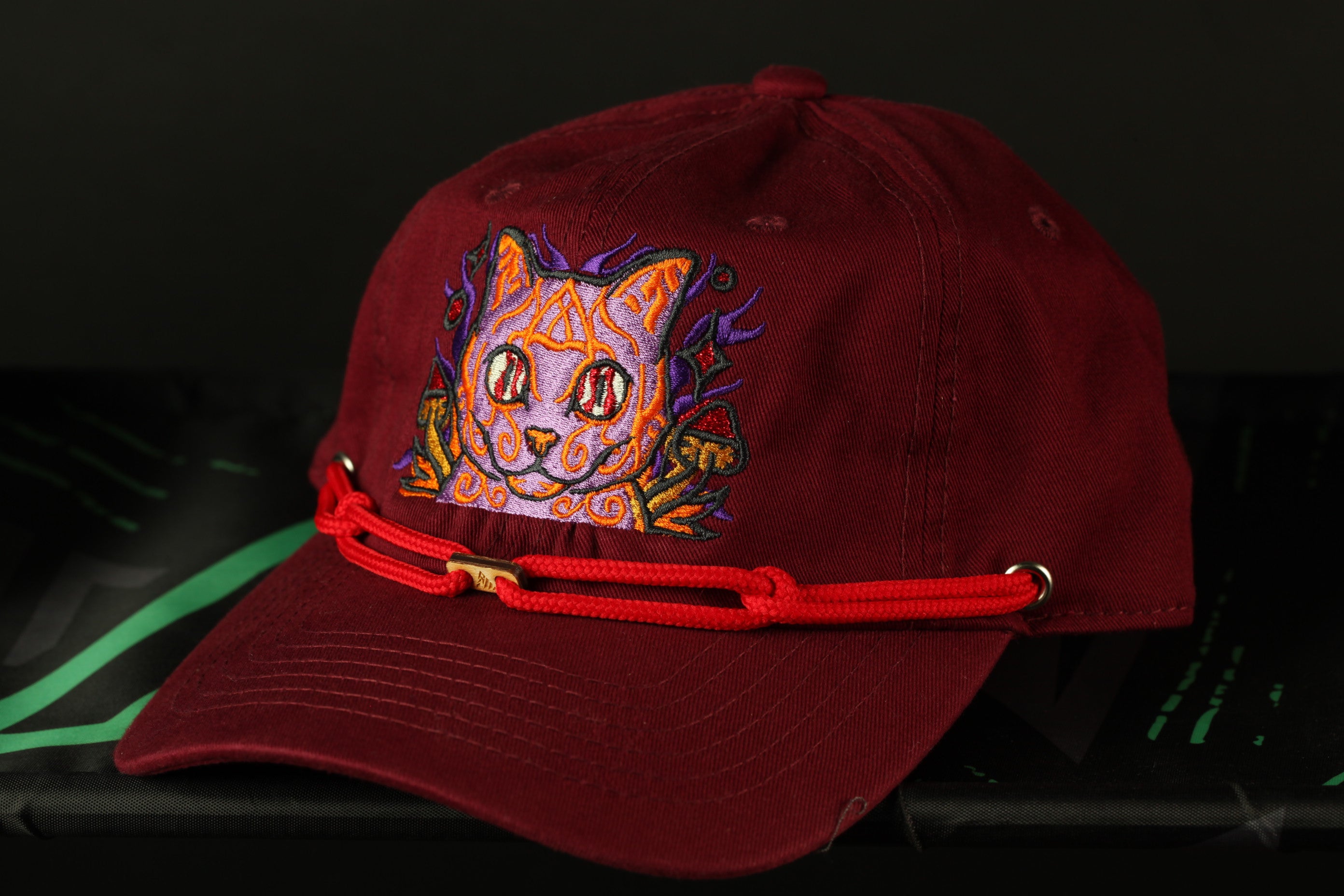 A burgundy baseball cap with a colorful cat embroidered on the front, featuring a red string or strap.
