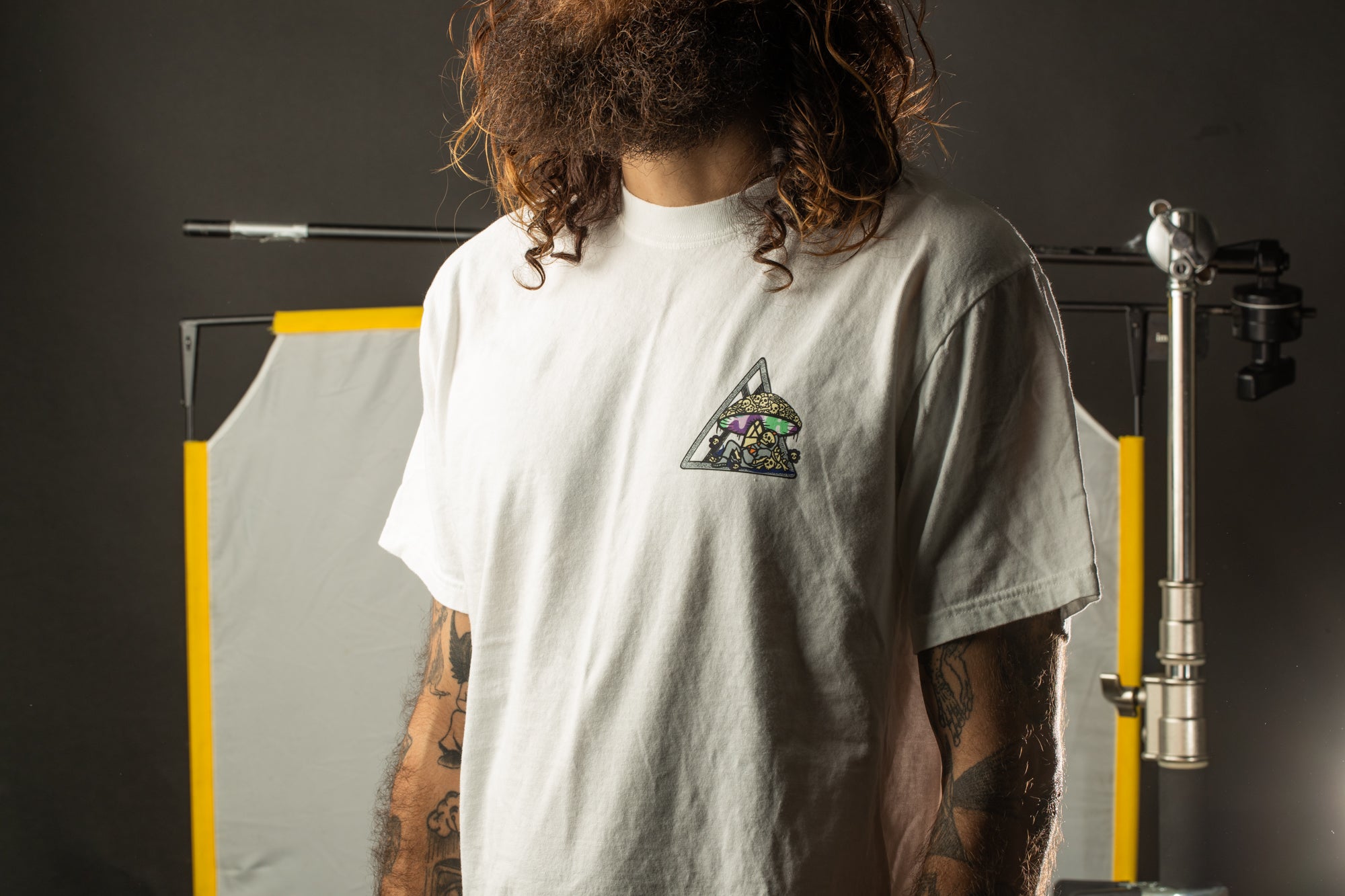 A person with long hair and tattoos is wearing a white t-shirt with a graphic design on it, standing in front of a dark background with a yellow and white striped object.