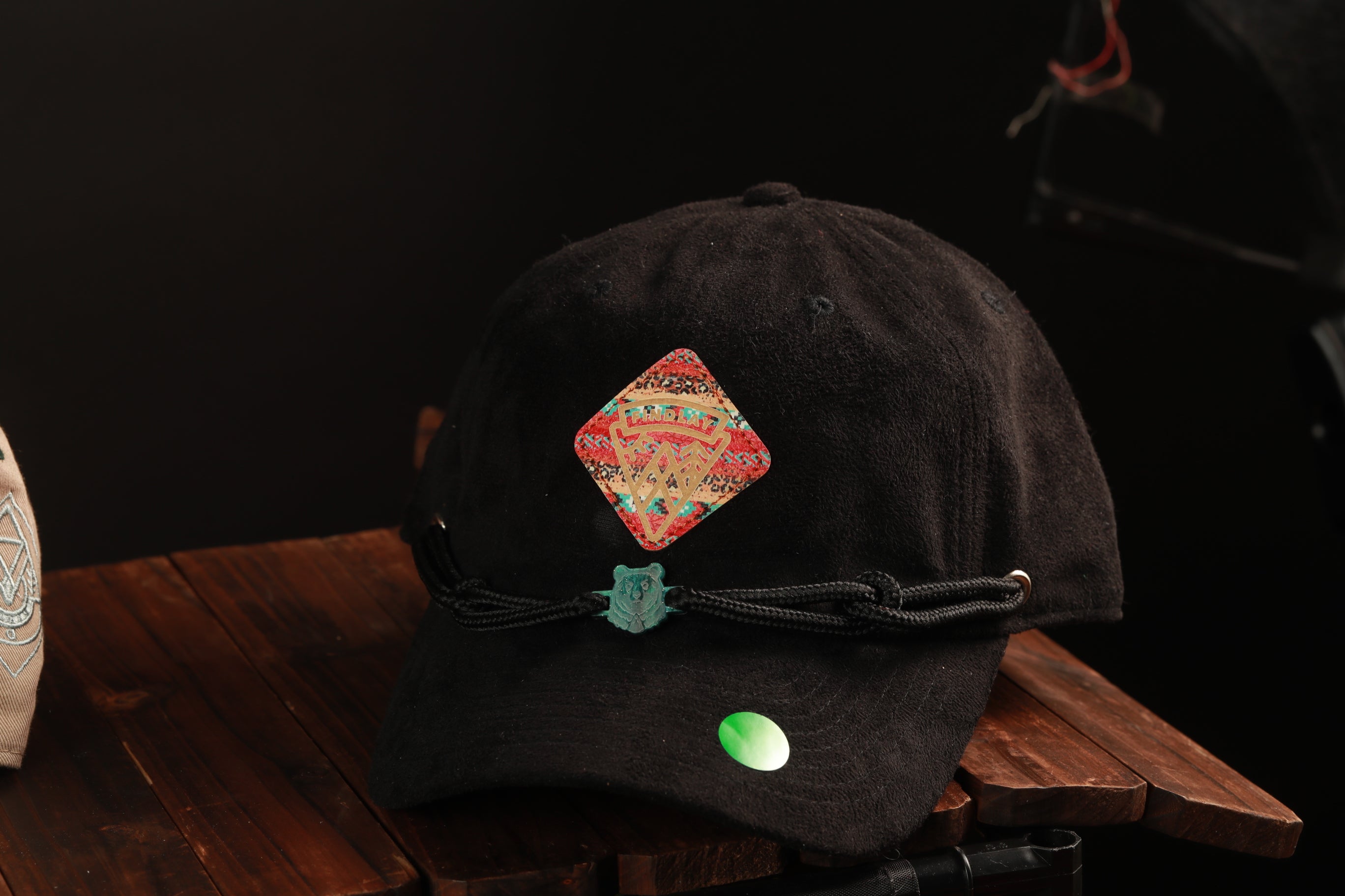 A black baseball cap with a colorful patch and a green button is displayed on a wooden surface.
