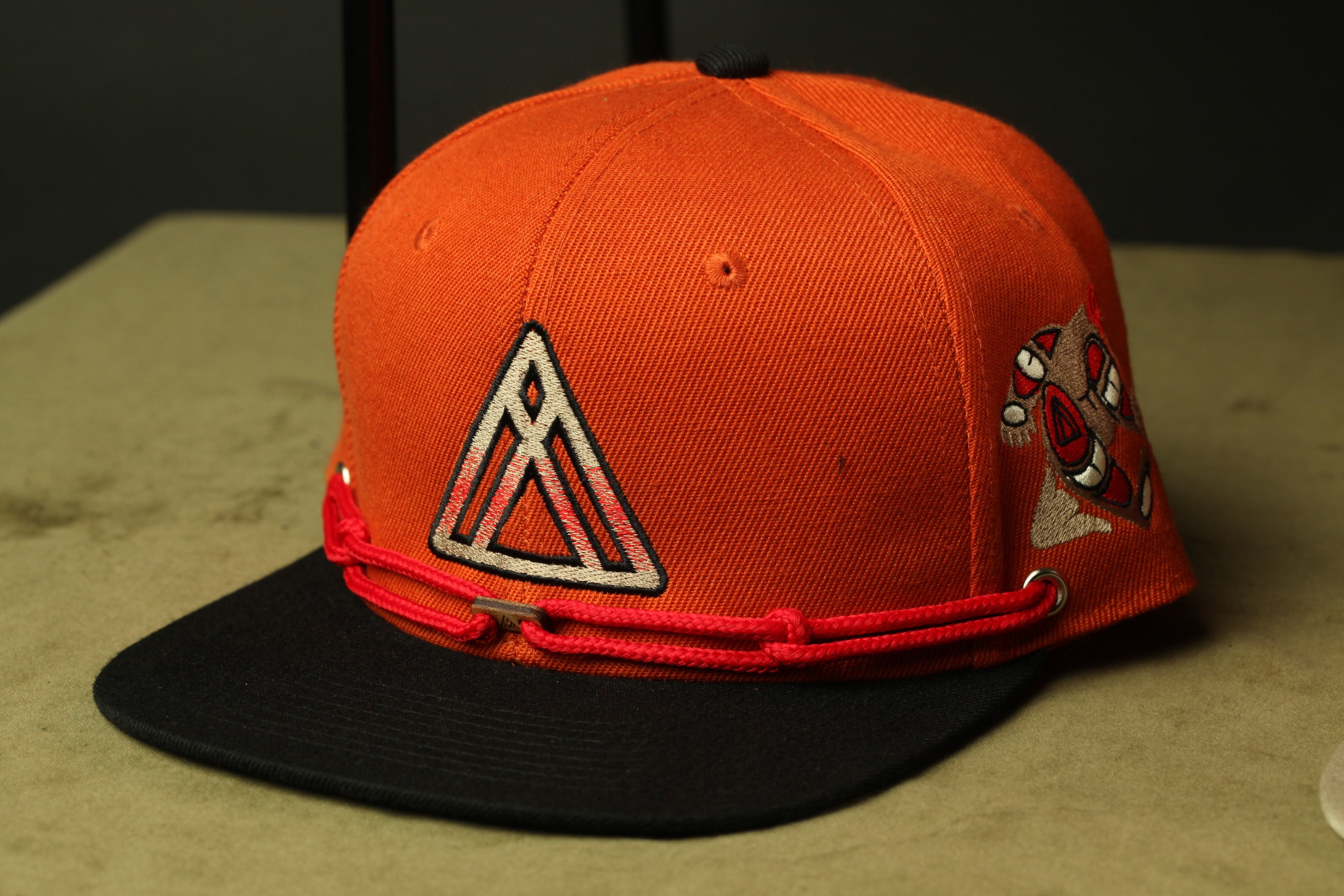 An orange baseball cap with a black brim and red straps, featuring a triangle logo on the front.