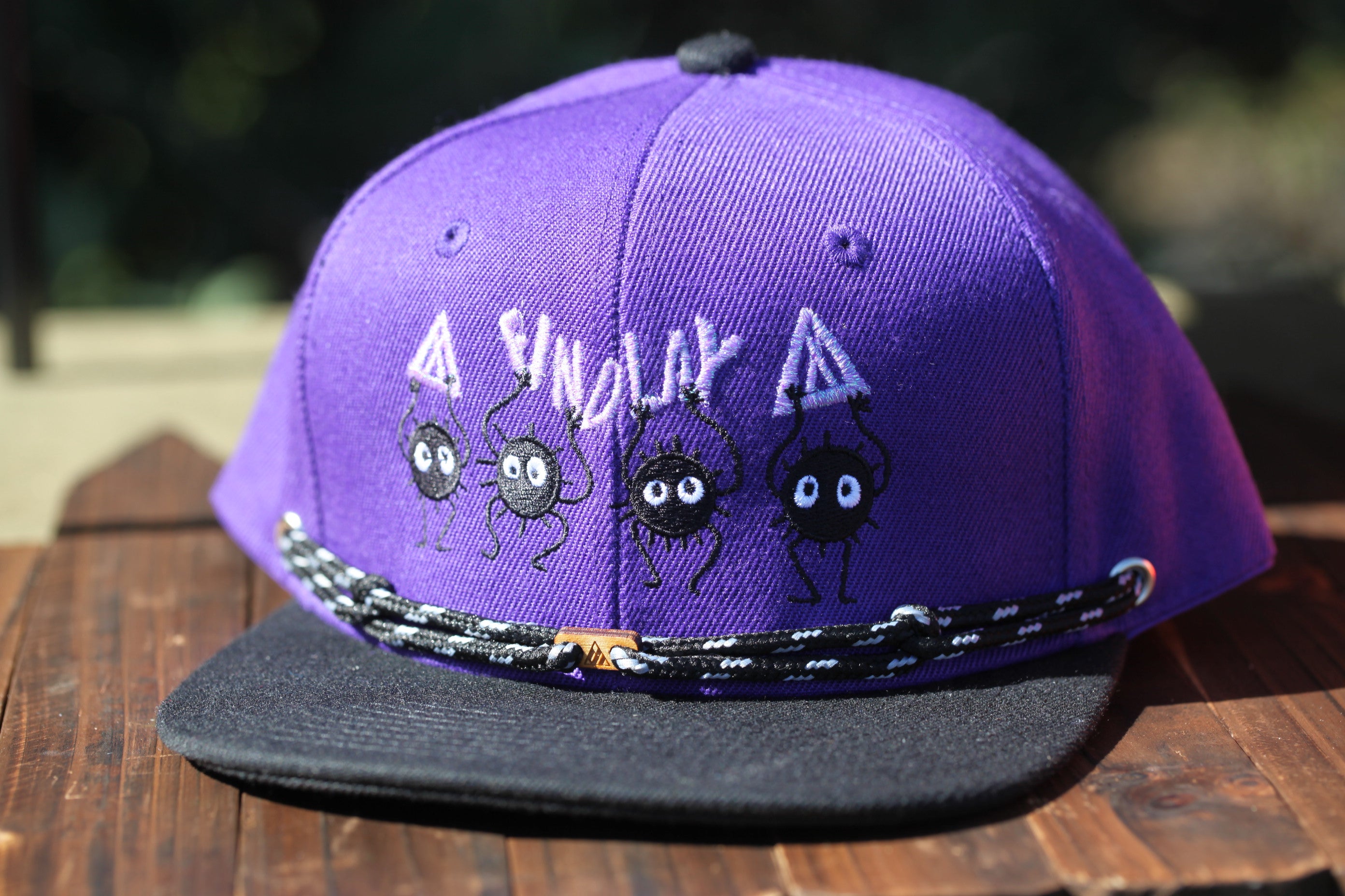 A purple baseball cap with a black brim and a black strap, featuring a playful design with cartoon-like black figures.