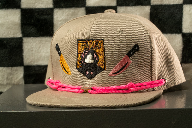 A beige baseball cap with a pink braided strap and various embroidered designs, including a shield-shaped emblem and a knife-like motif.