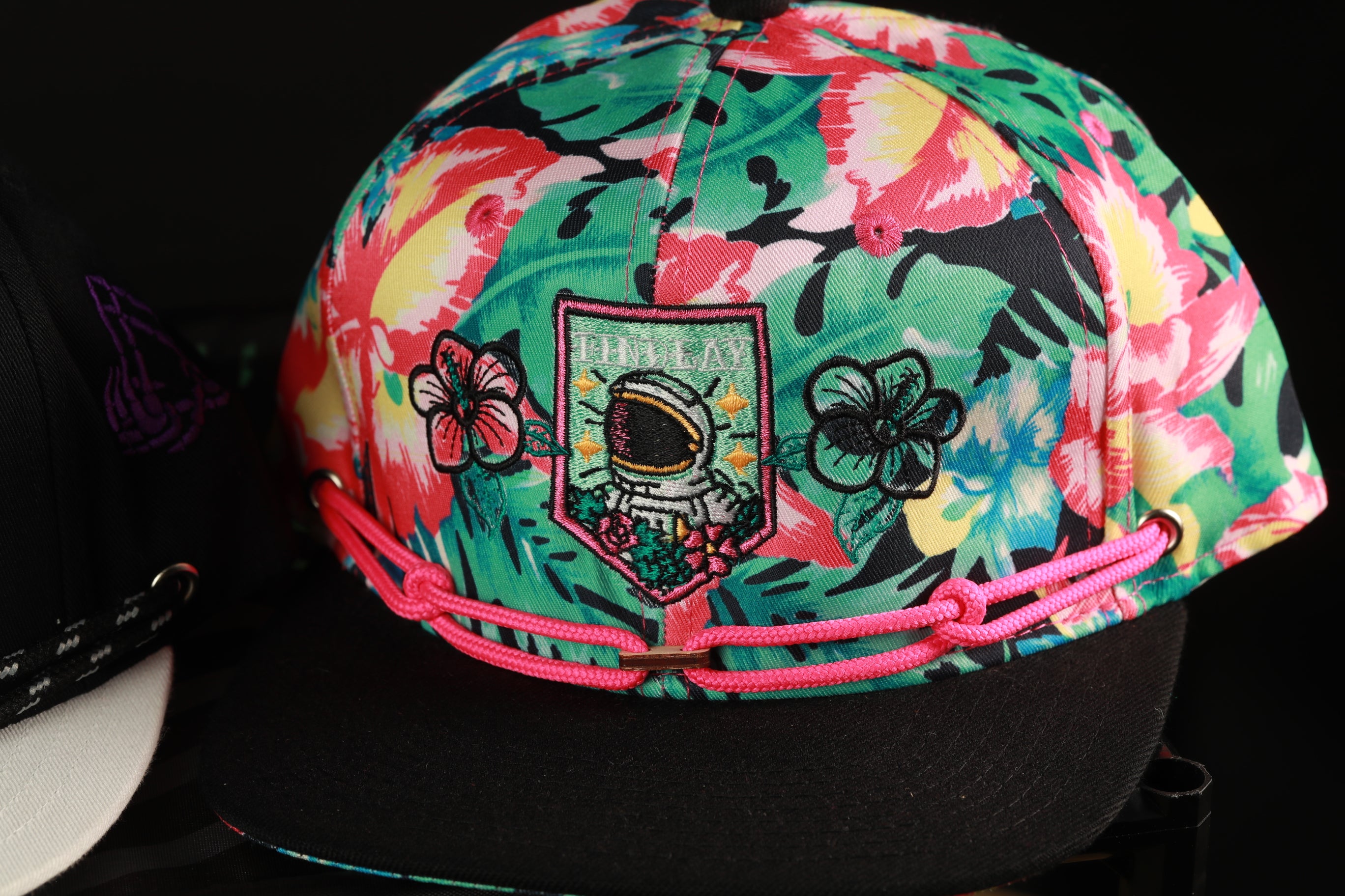 A colorful floral-patterned baseball cap with a pink braided strap and a small embroidered patch on the front.