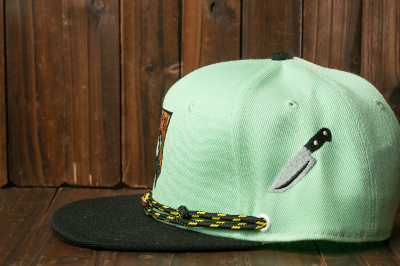 A light green baseball cap with a black brim and a small knife embroidered on the side, placed on a wooden surface.