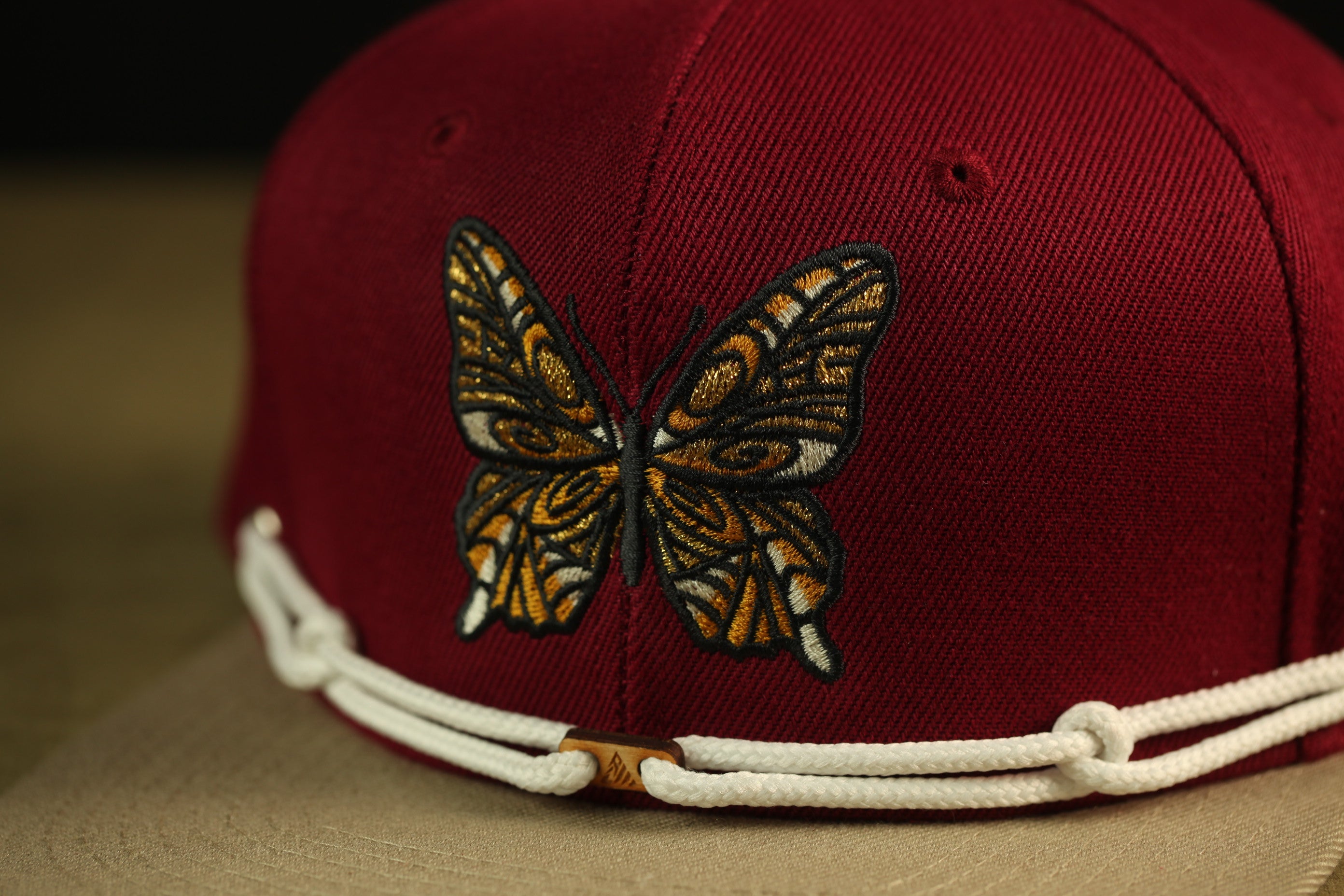 A close-up view of a maroon baseball cap with a detailed embroidered butterfly design on the front.