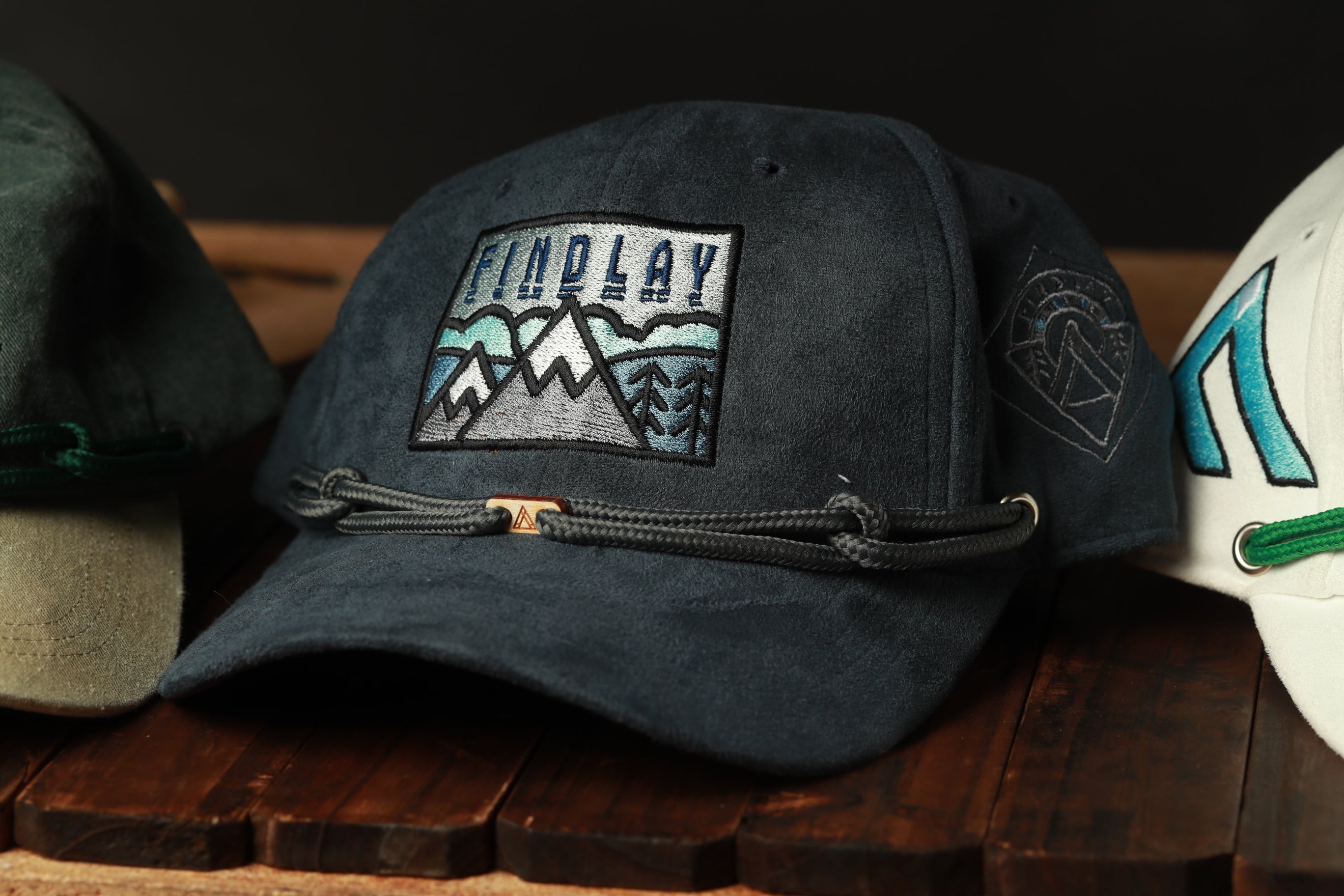 A close-up image of a black baseball cap with a mountain design and the word "FINDlay" printed on it, placed on a wooden surface.