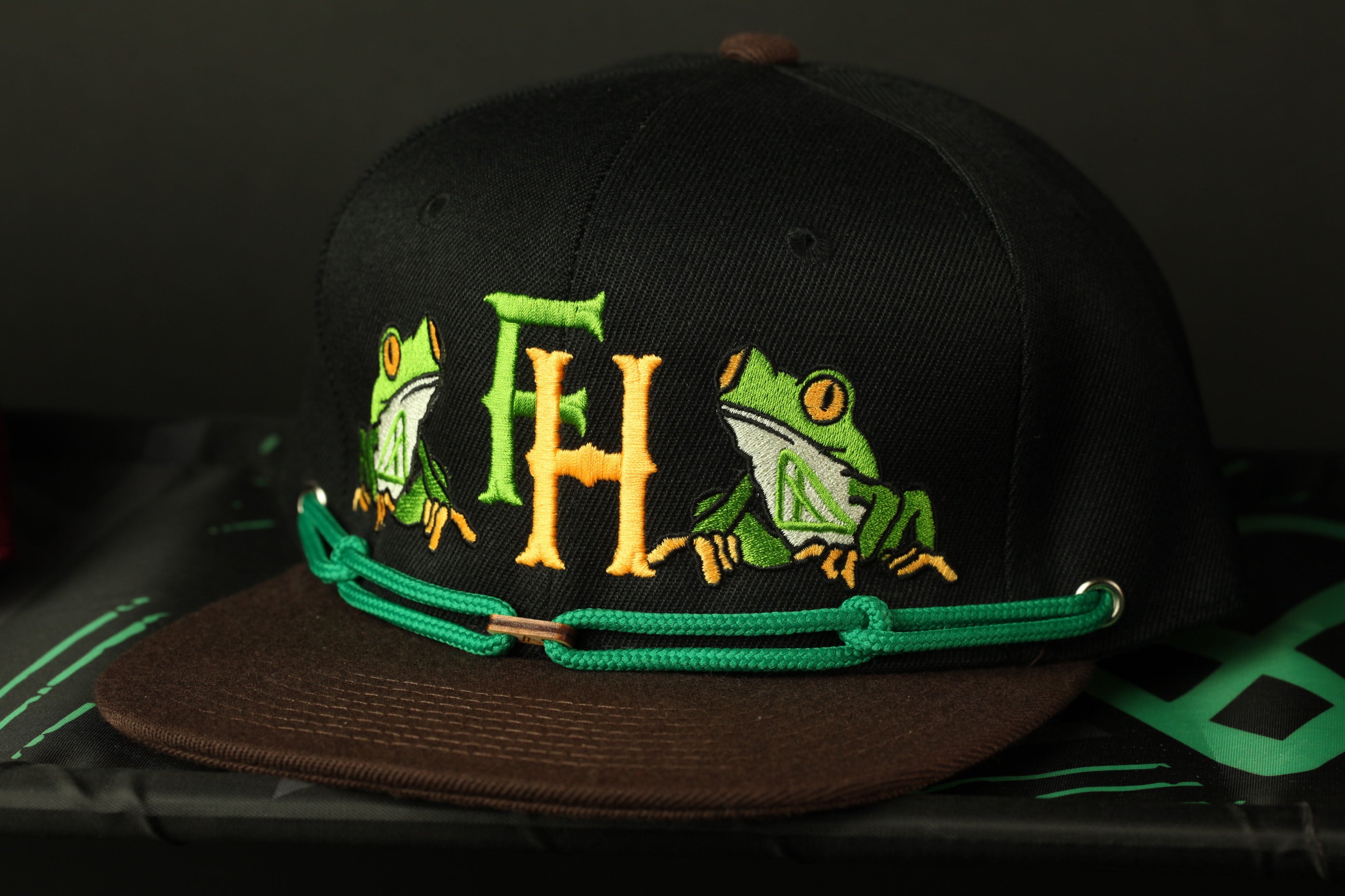 A black baseball cap with a green and yellow frog embroidered on it, sitting on a black surface.