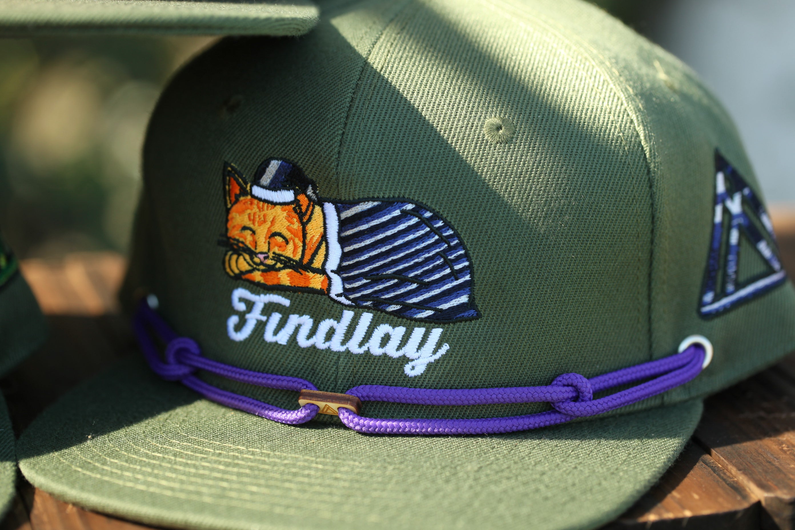 A close-up image of a green baseball cap with a cartoon cat design and the word "Findlay" printed on it.