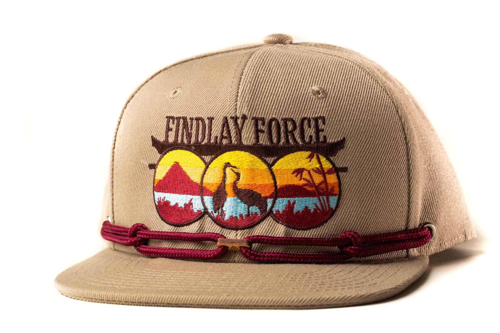 A tan baseball cap with a maroon band and a graphic design featuring a bird silhouette and a sunset landscape.
