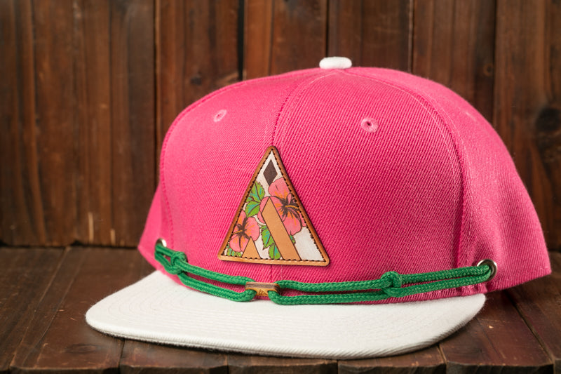 A pink baseball cap with a floral patch and green string is displayed against a wooden background.
