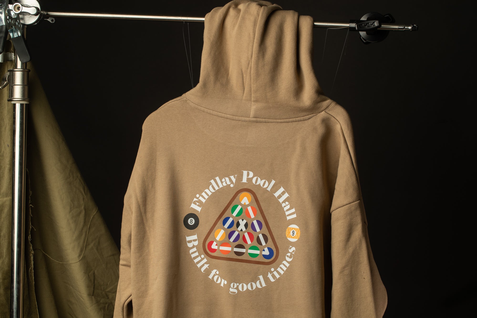 A beige hoodie with a pool-themed design and text printed on it, hanging on a metal hanger against a dark background.