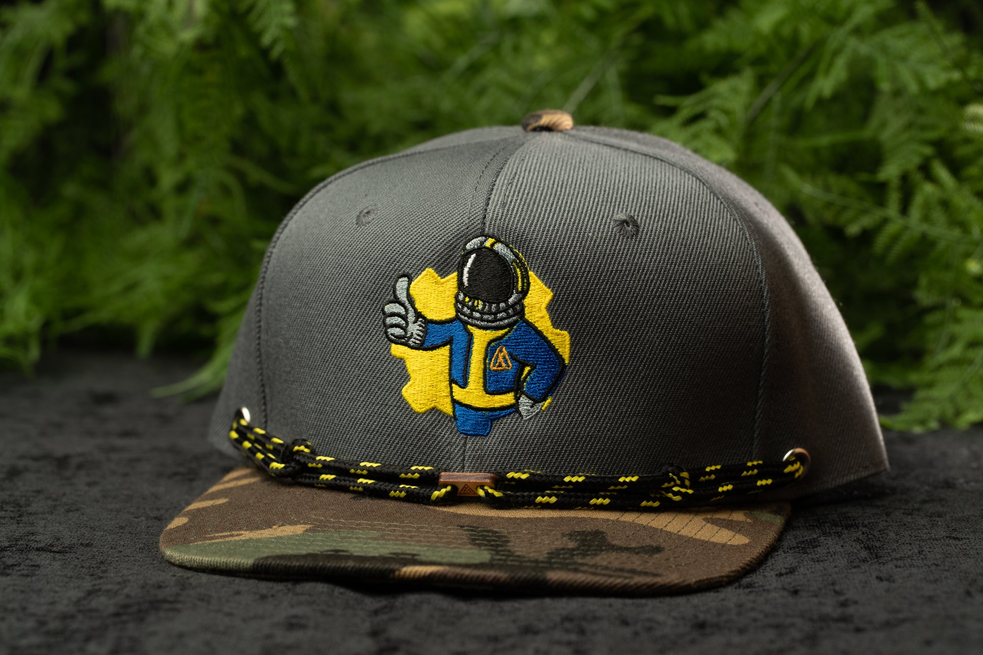 A gray baseball cap with a yellow and blue embroidered character on it, sitting on a camouflage patterned brim.