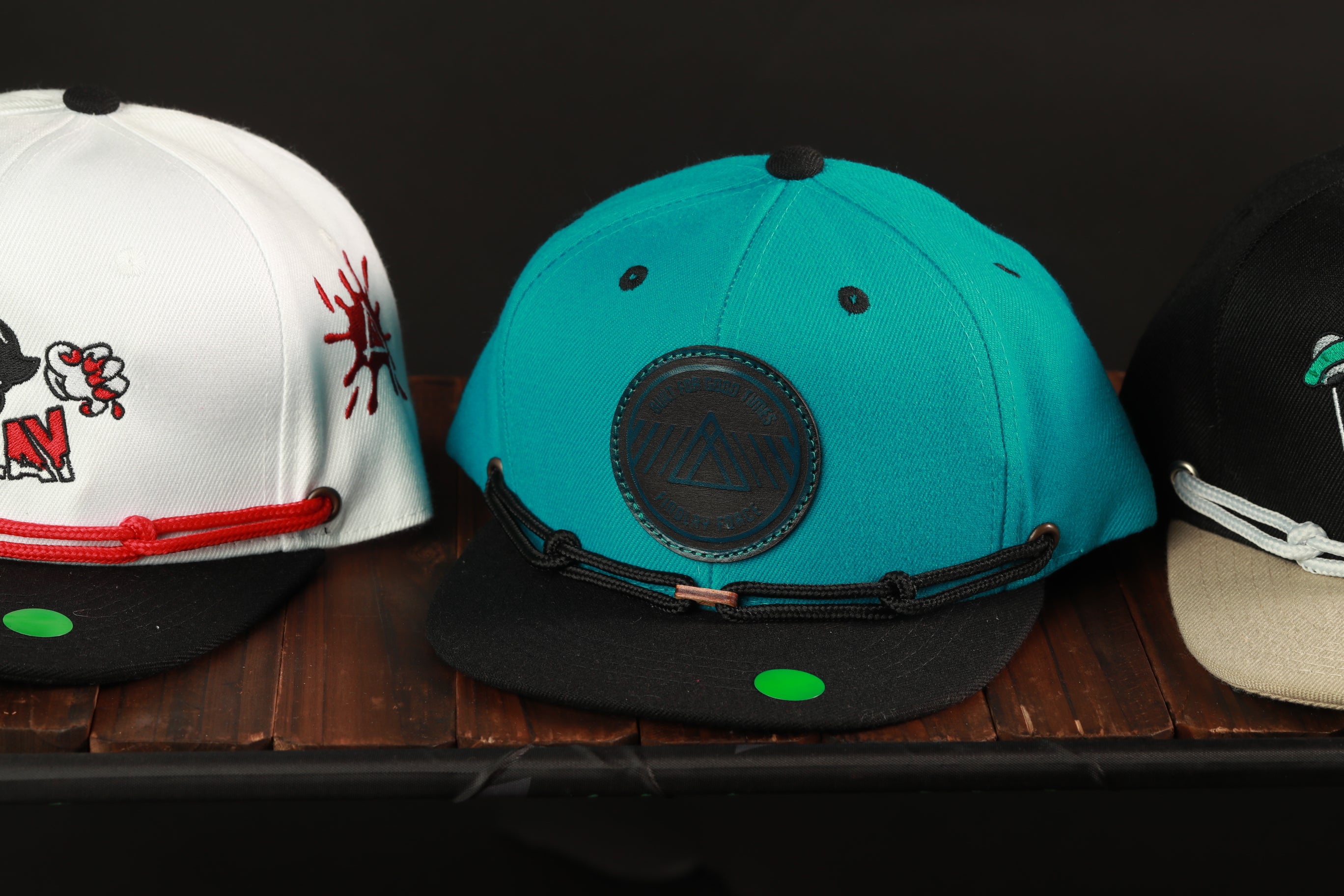 Three baseball caps are displayed on a wooden surface, each with distinct designs and colors.