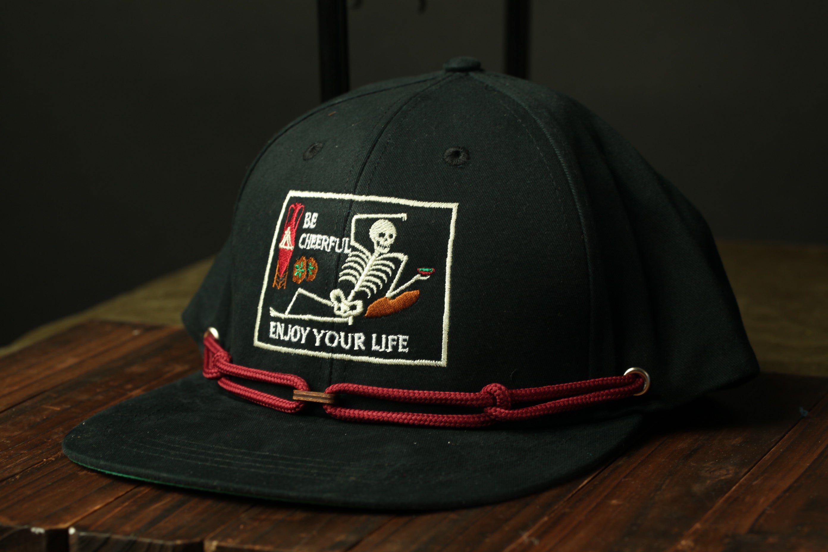 A black baseball cap with a red string and a white patch on the front, featuring a skeleton and the text "Be Cheerful, Enjoy Your Life".