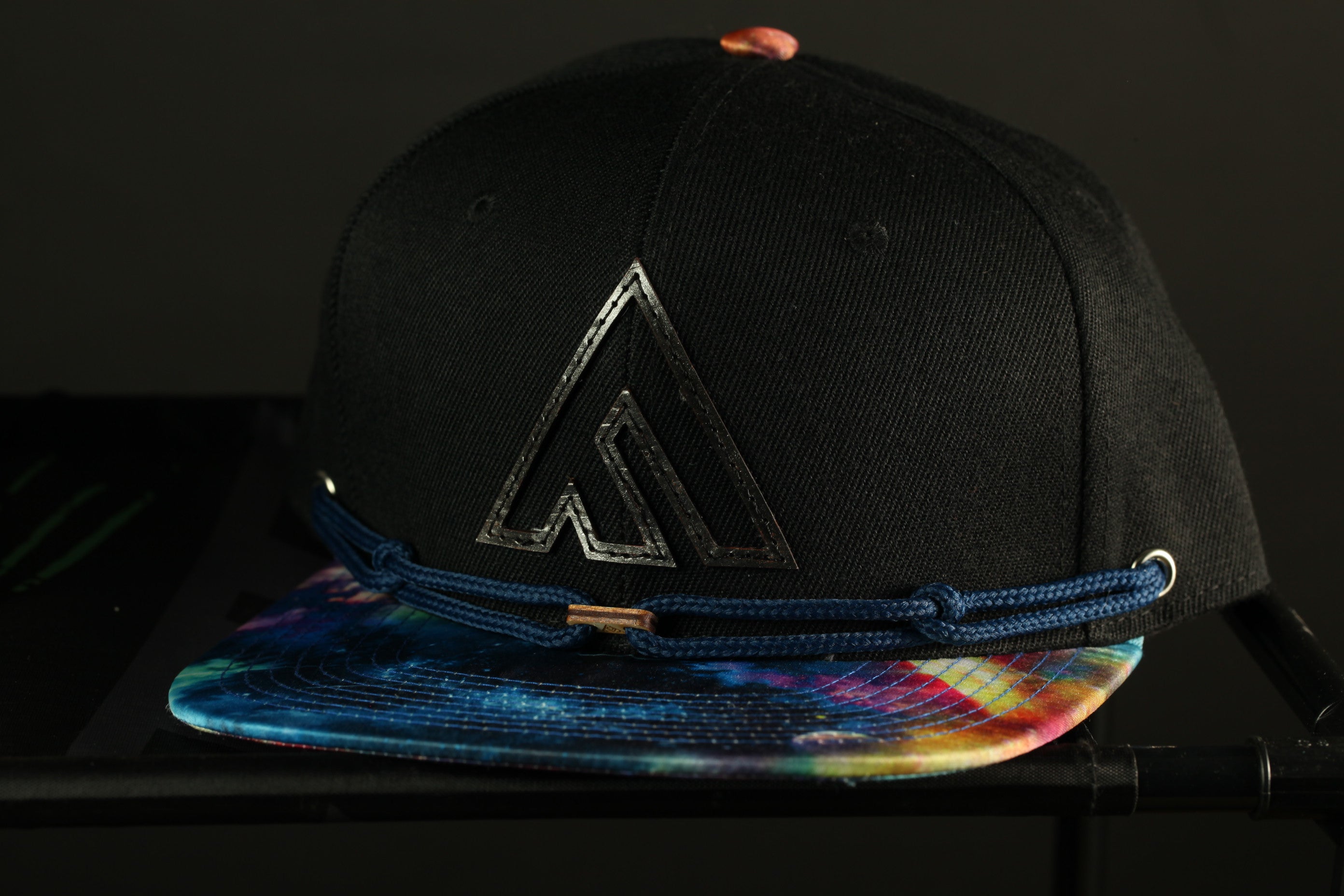 A black baseball cap with a tie-dye pattern and a logo on the front, placed on a black surface.