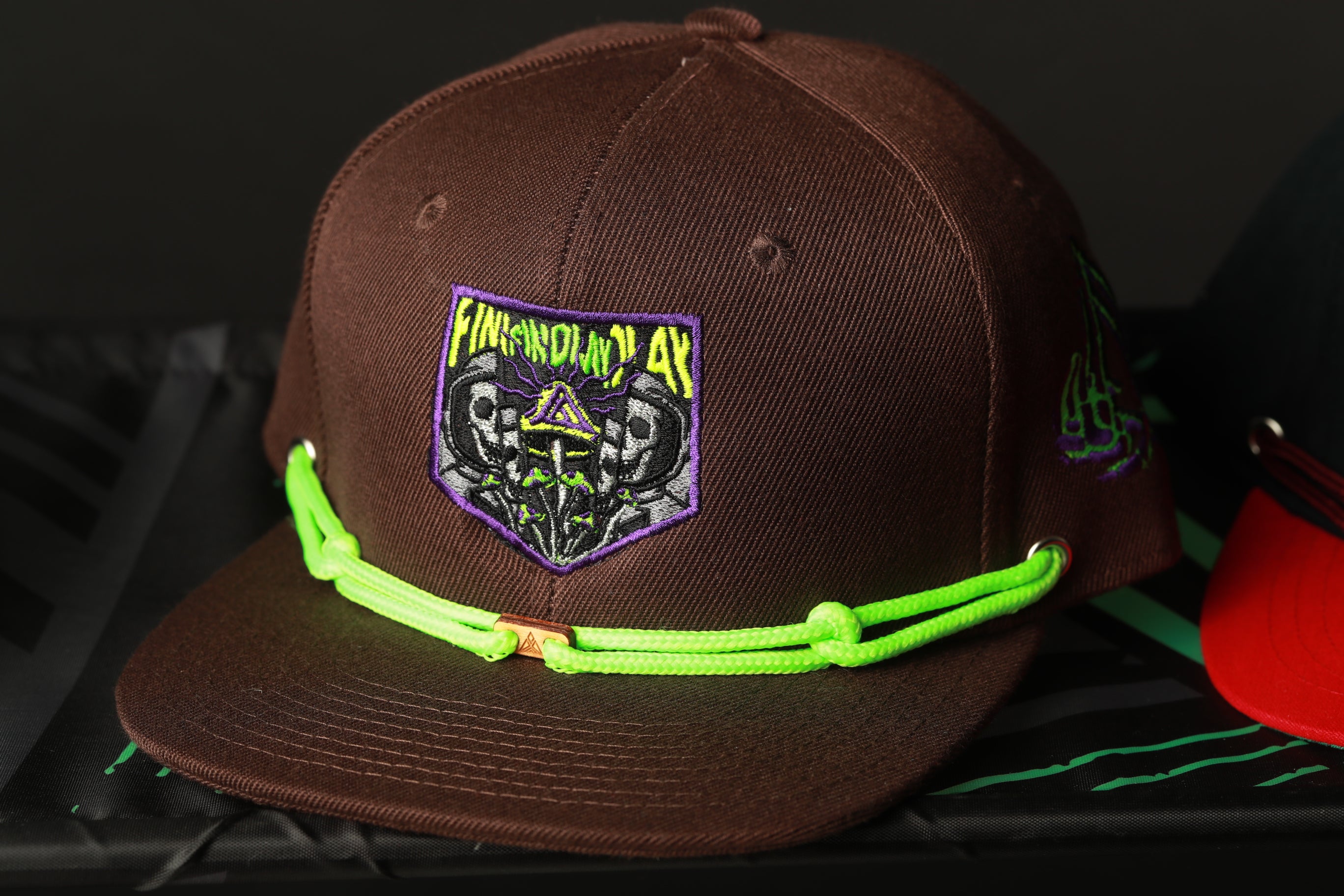 A brown baseball cap with a green string and a purple and green patch featuring a skull and crossbones design.