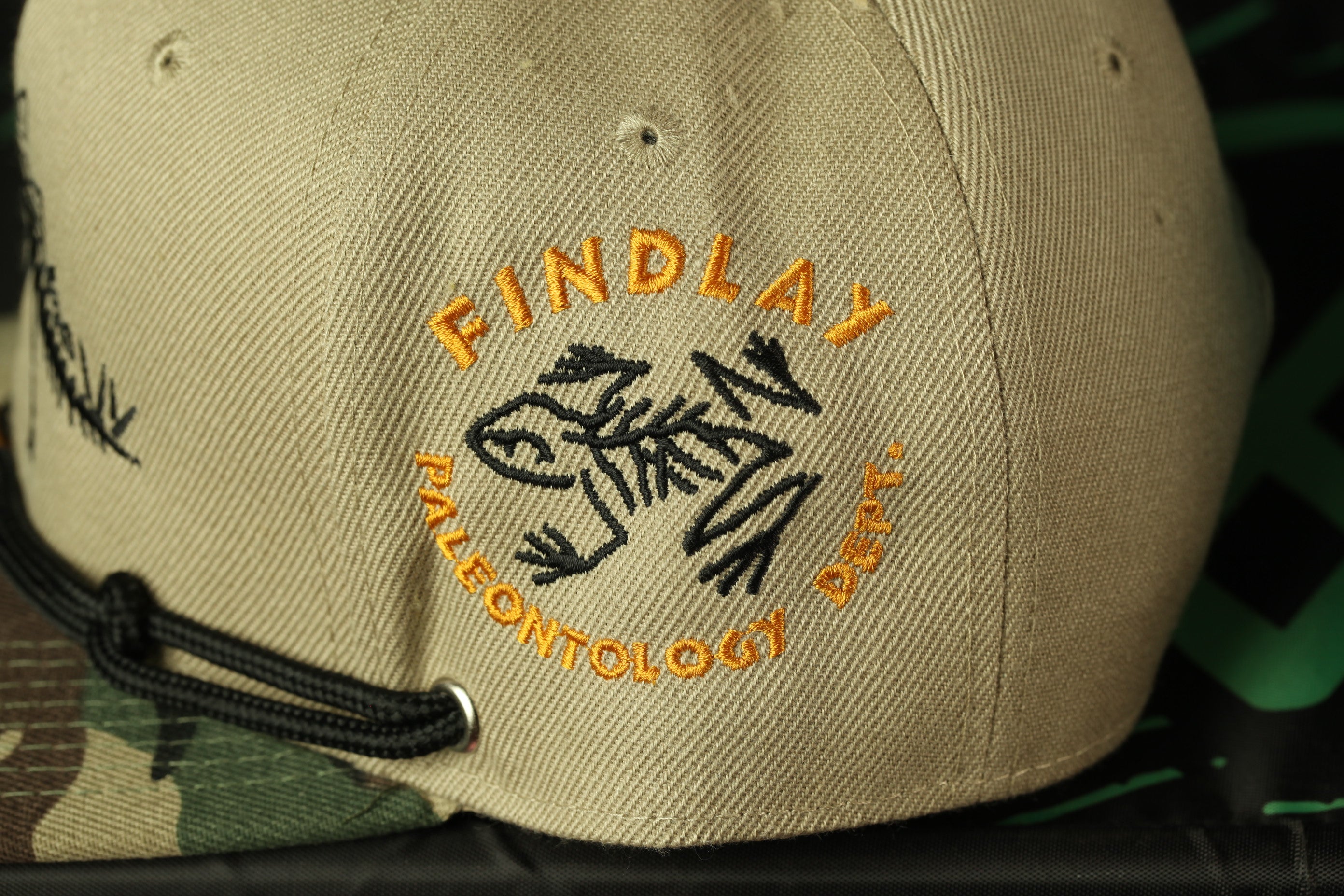 A beige baseball cap with a black and orange logo on the front.