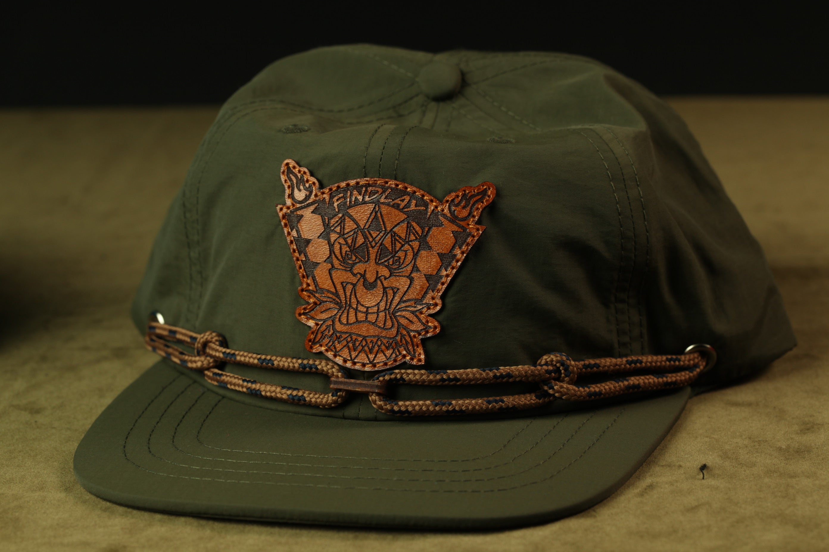 A green baseball cap with a leather patch featuring a stylized design on the front.