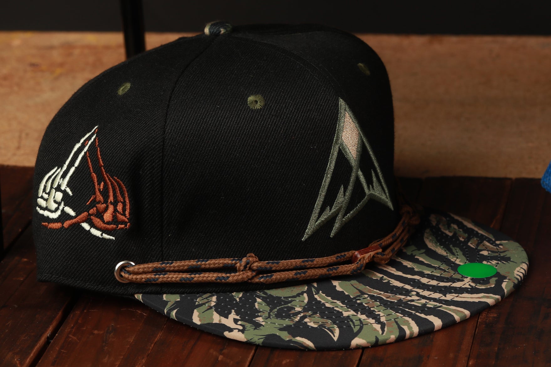 A black baseball cap with a camouflage pattern and a graphic design on the front, placed on a wooden surface.