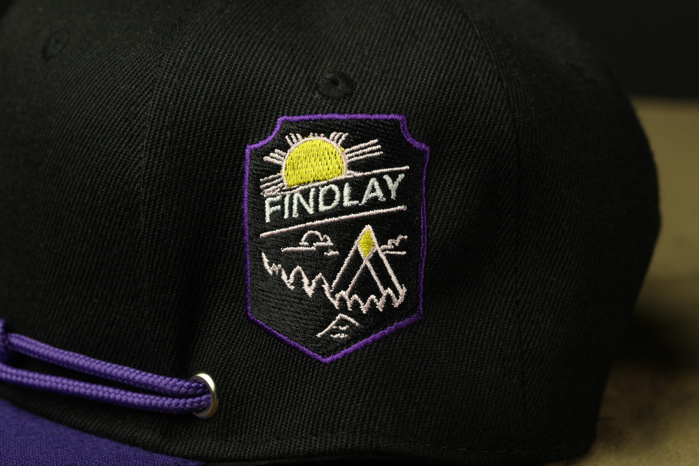 A close-up view of a black baseball cap with a purple and yellow embroidered logo on the front.
