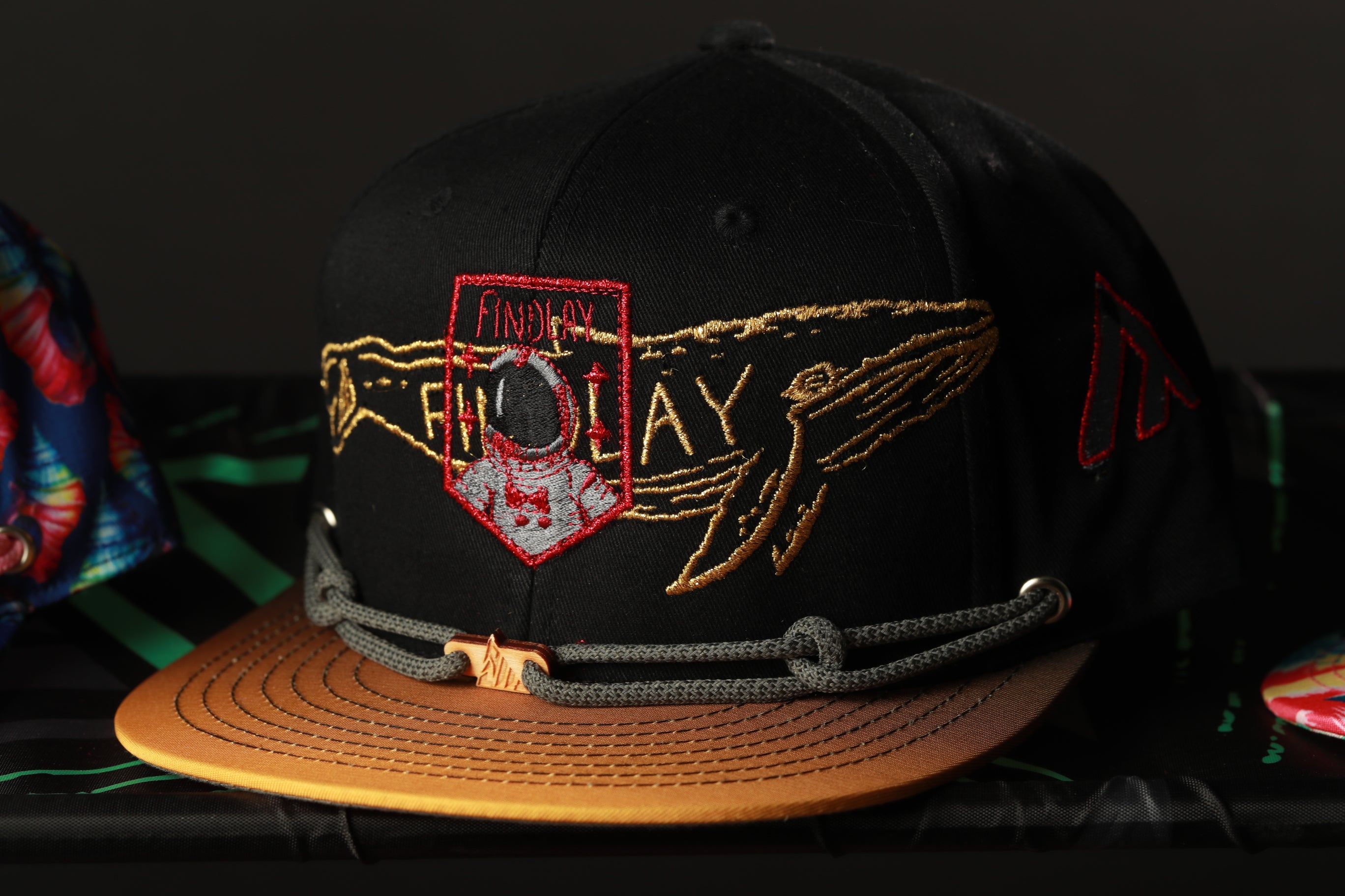 A black baseball cap with a gold and red embroidered design, featuring a whale and the text "FINDA" on the front.