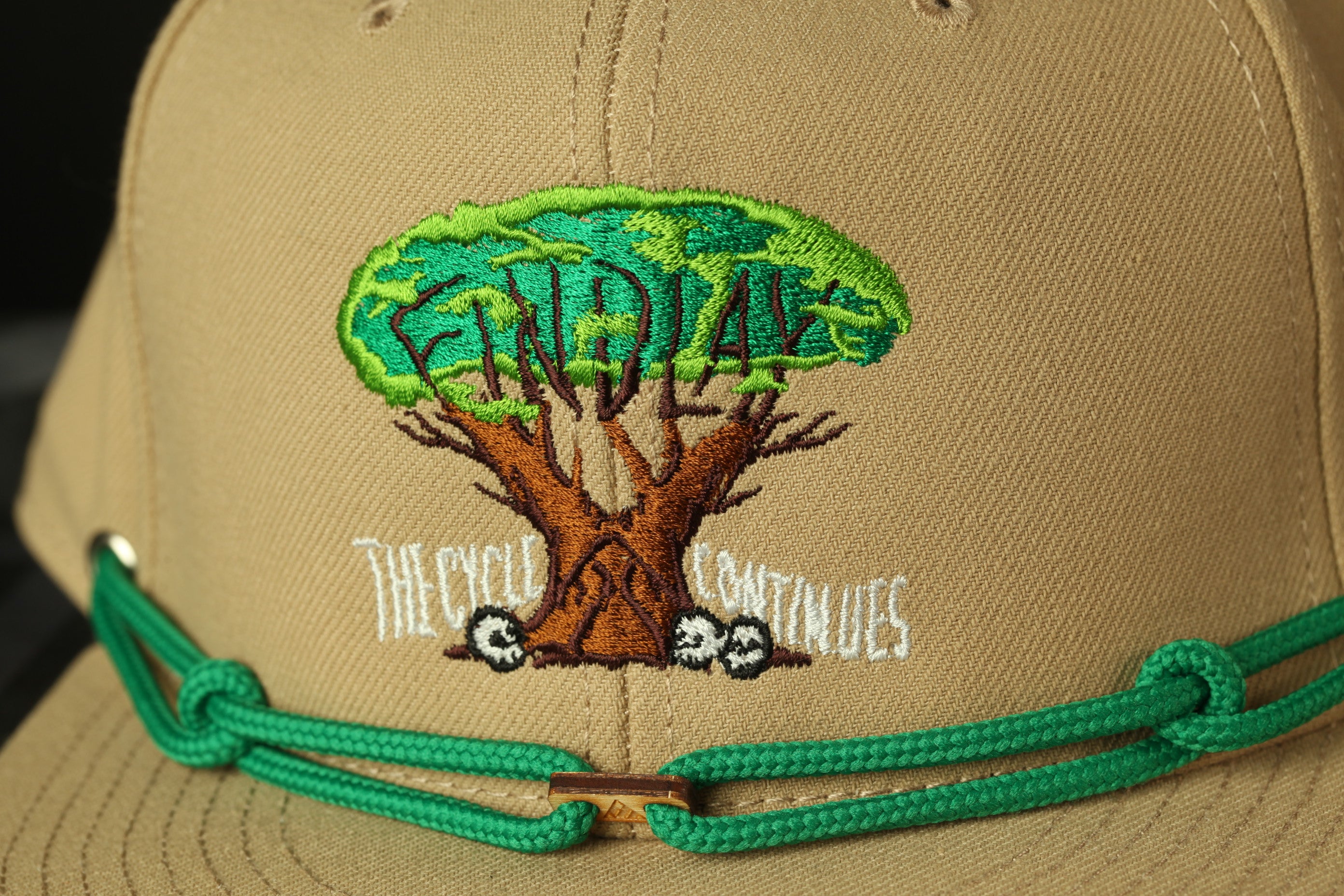 A close-up of a beige baseball cap with a green string and a tree embroidered on it.