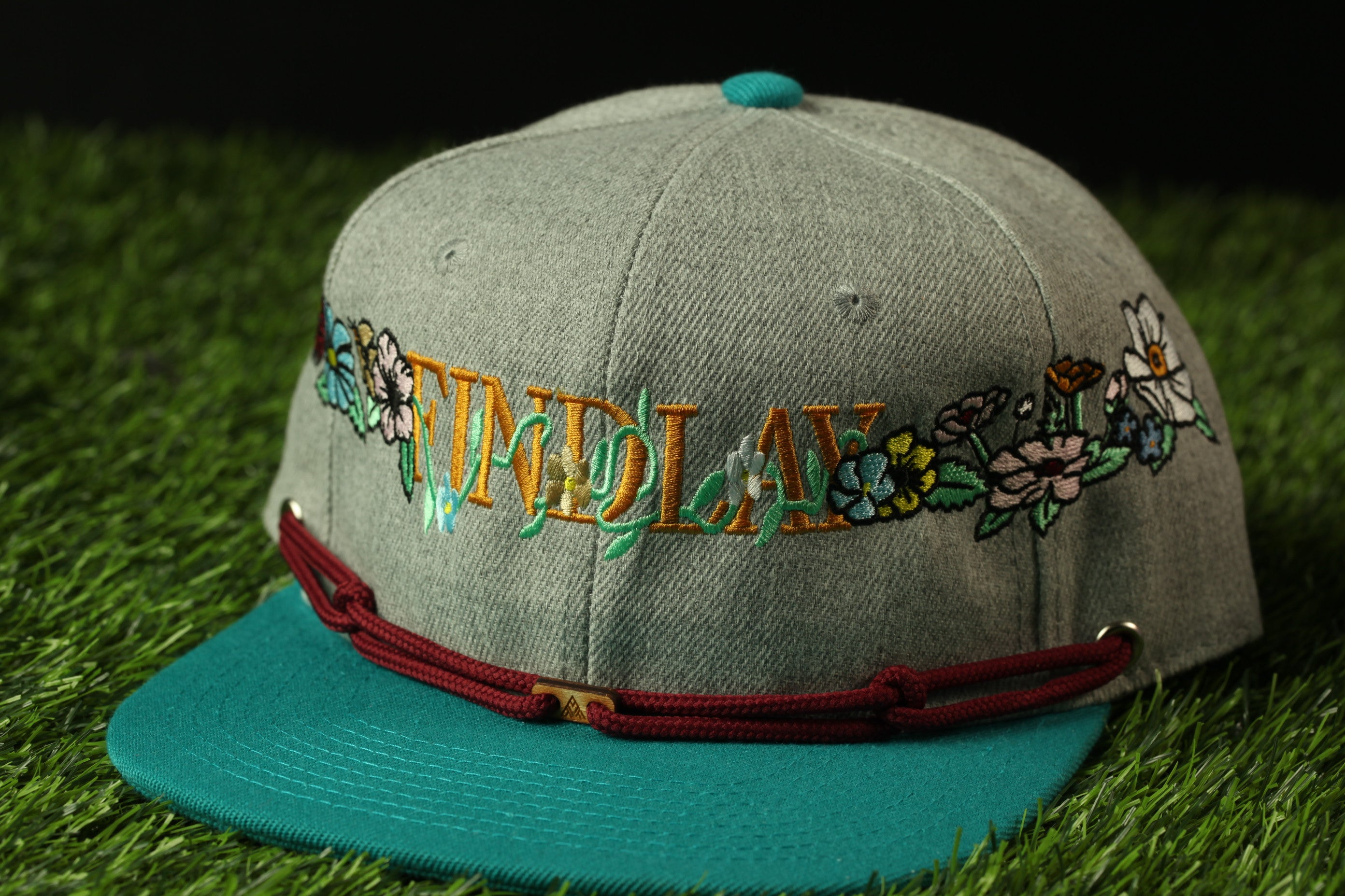 A gray baseball cap with a teal brim and a red string, featuring embroidered text and colorful floral designs.