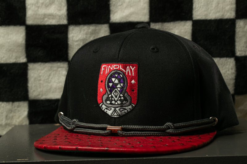 A black baseball cap with a red and black patterned brim and a red and black logo on the front, placed on a black surface against a black and white checkered background.