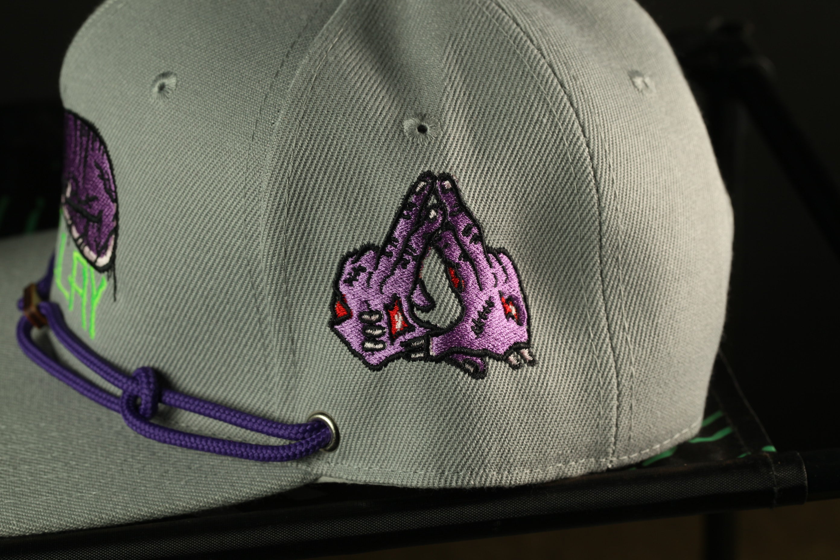 A close-up view of a gray baseball cap with purple and green embroidered designs, including a graphic of a hand with a skull-like design.