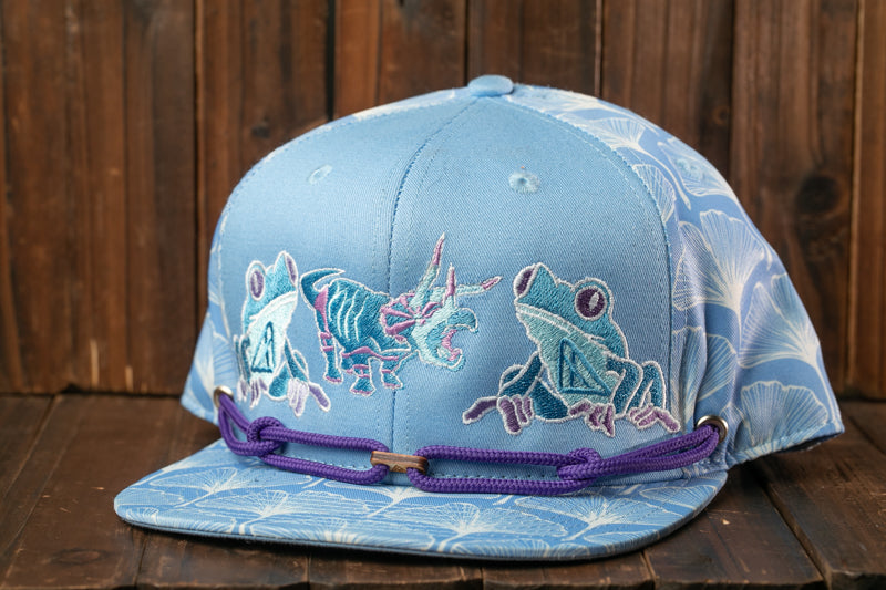 A blue baseball cap with a purple string and a design of three cartoon frogs on the front.