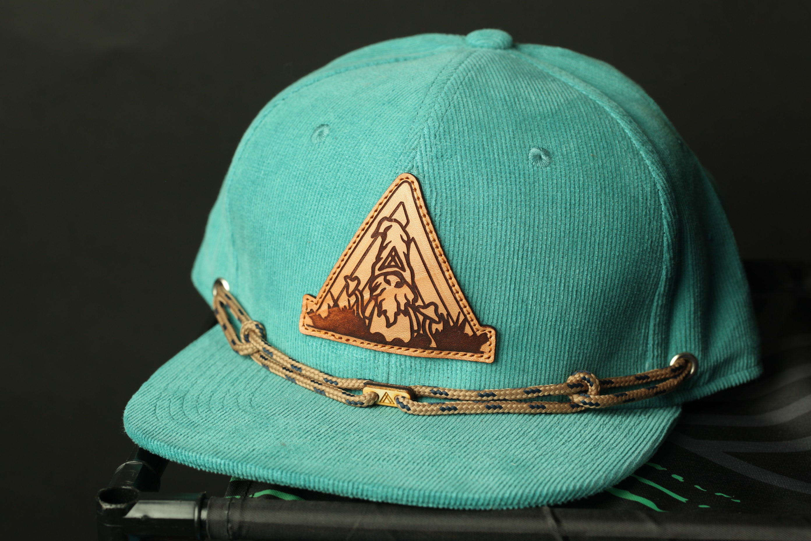 A turquoise baseball cap with a leather patch featuring a mountain landscape design.
