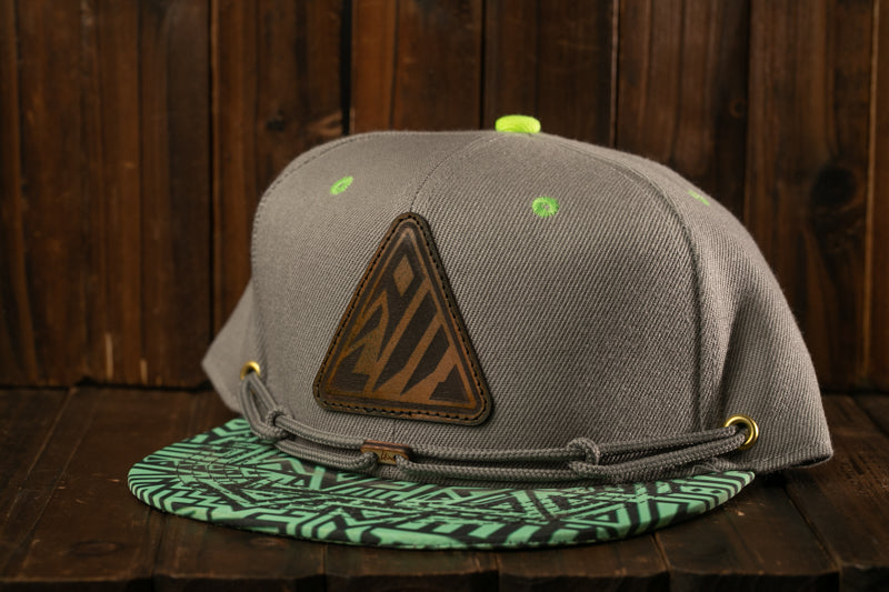 A gray baseball cap with a green geometric pattern on the front and a leather patch on the brim, placed on a wooden surface.