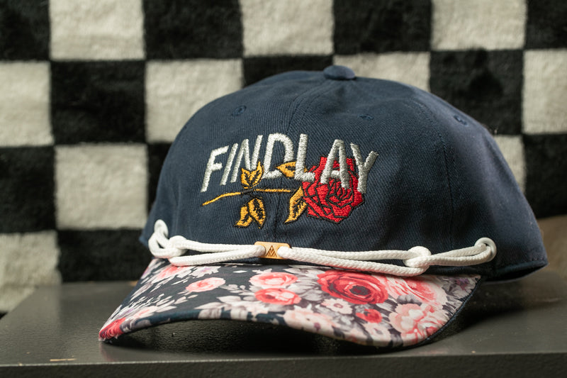 A navy blue baseball cap with a floral pattern and the word "FINDlay" embroidered on it, sitting on a table against a black and white checkered background.